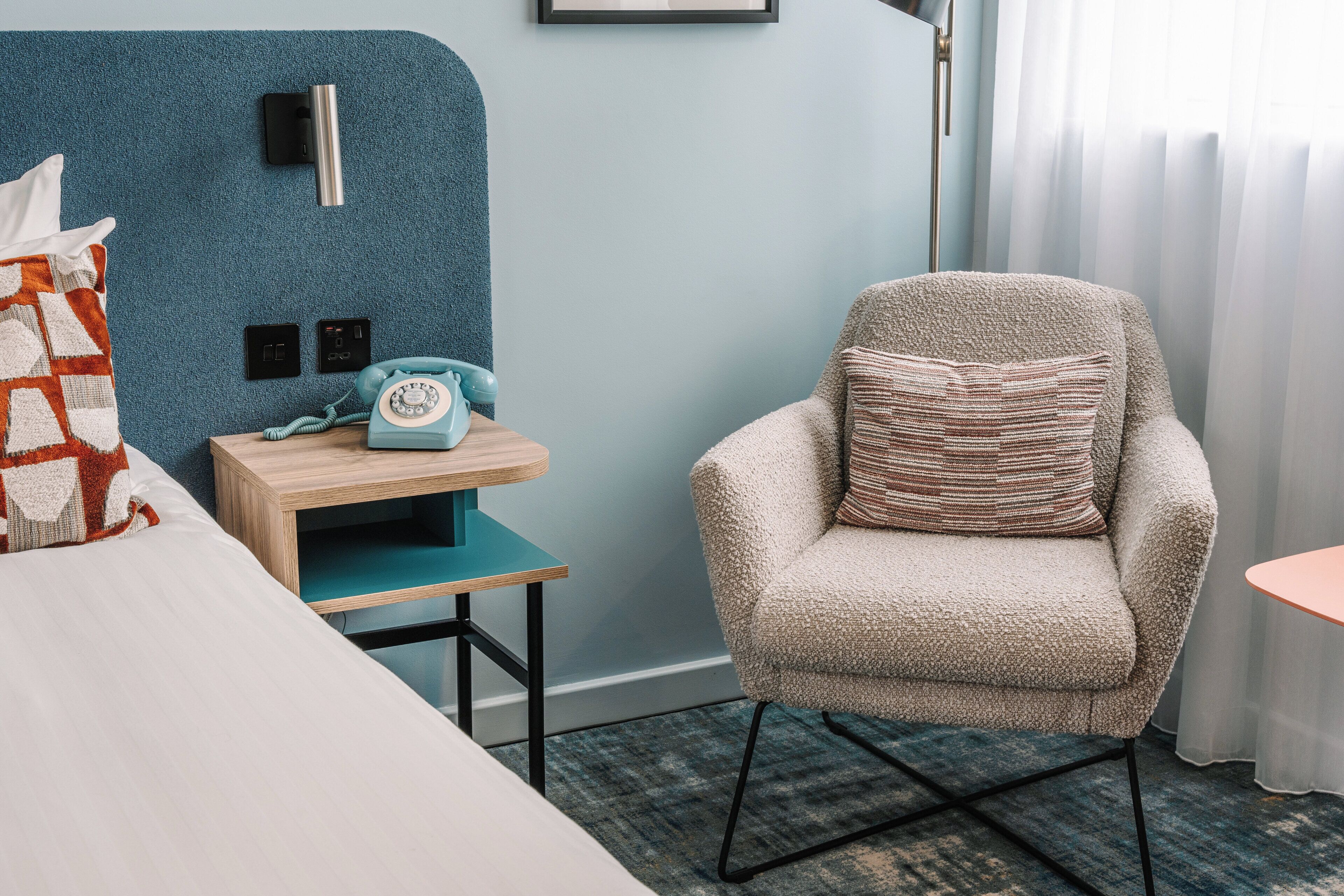 A hotel room with a bed, a chair, a small table, a phone, and a wall-mounted light.