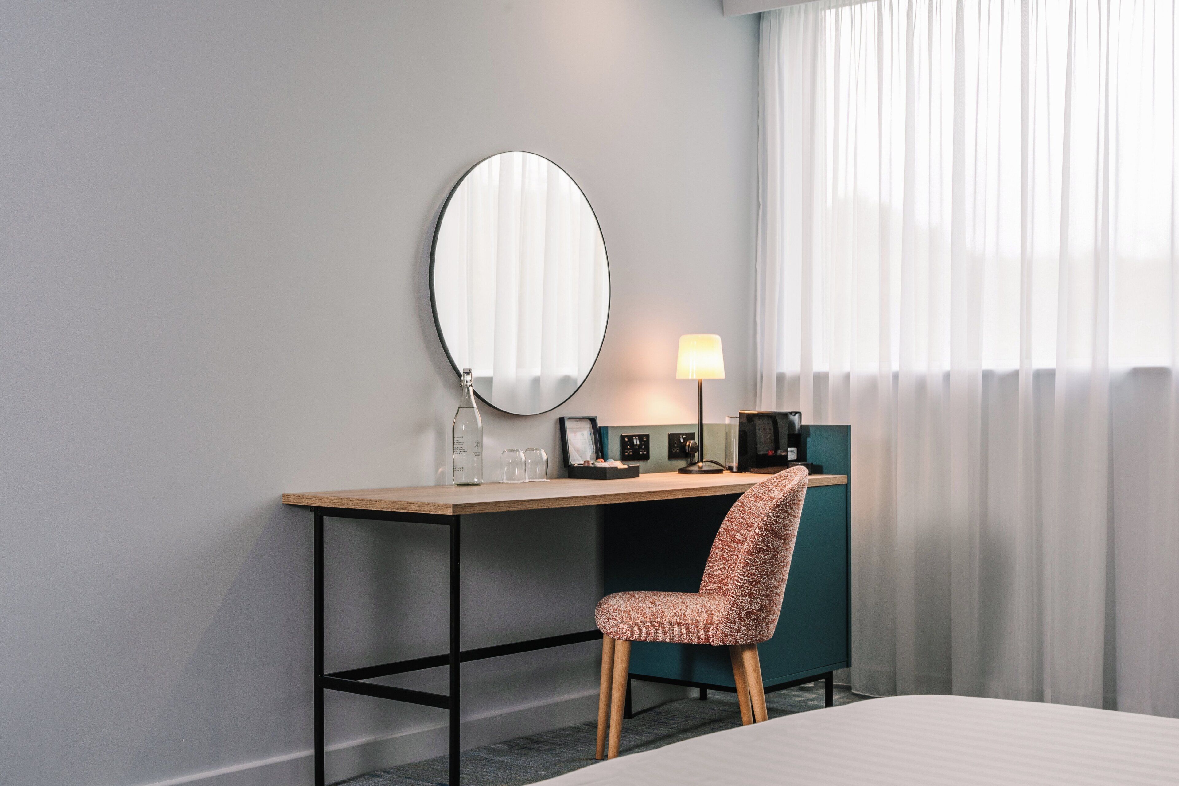 Une chambre d’hôtel avec un bureau en bois, un miroir rond, une chaise, une lampe et un lit.