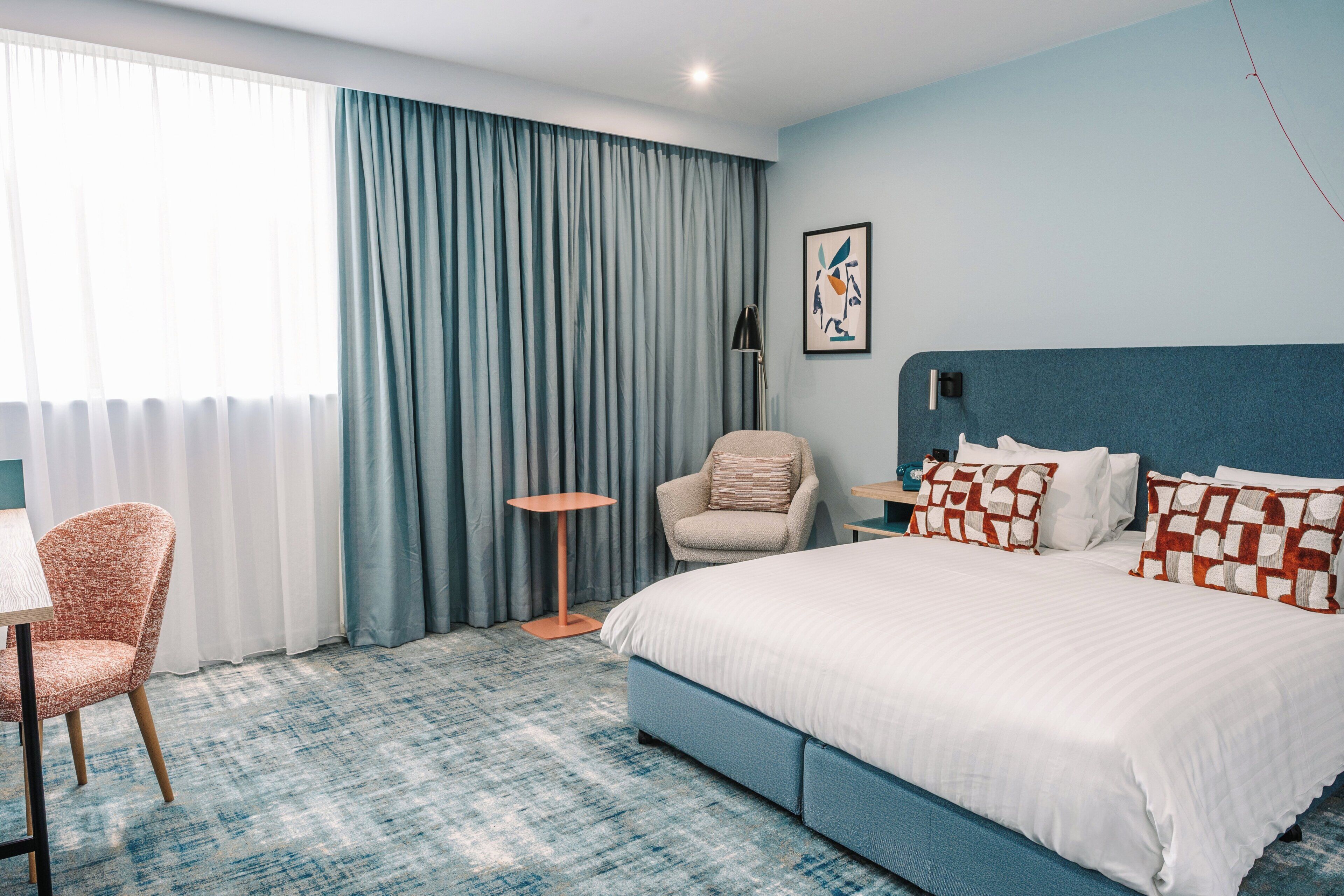 Quarto de hotel com uma cama grande, uma cadeira, uma mesinha e um abajur.