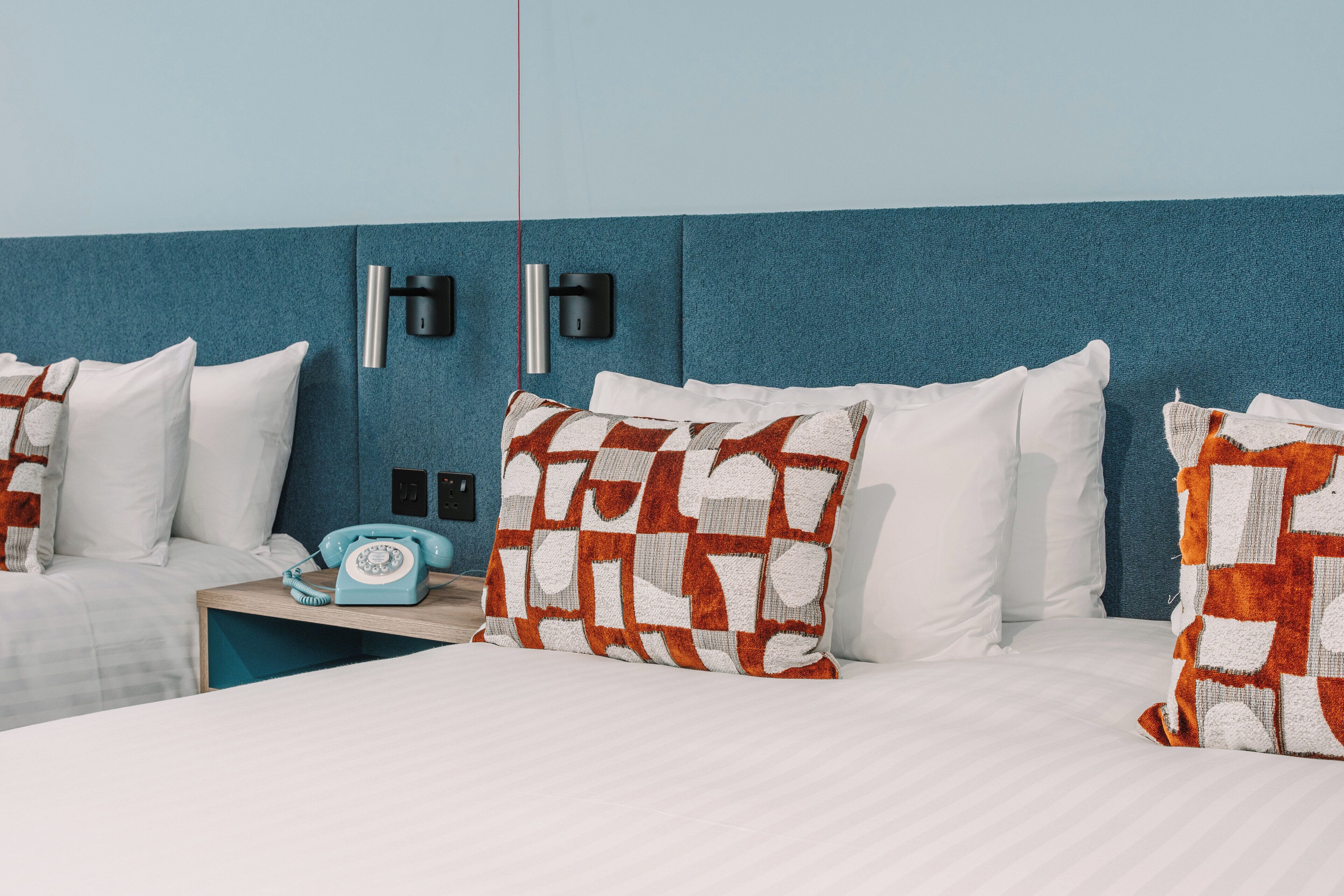 A hotel room with a blue accent wall, two beds, white bedding, patterned pillows, a vintage-style telephone, and wall-mounted light fixtures.