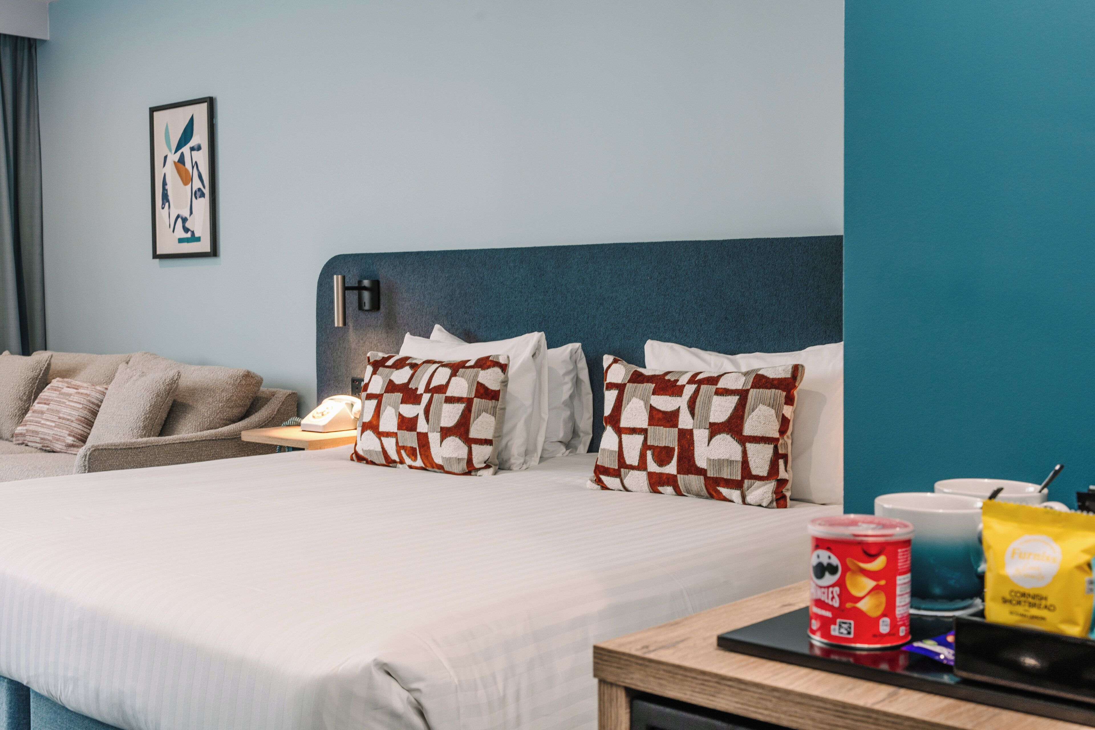 Une chambre d’hôtel avec un grand lit, deux oreillers à motifs à carreaux, une table de chevet avec une lampe, un téléphone et un plateau avec des en-cas.
