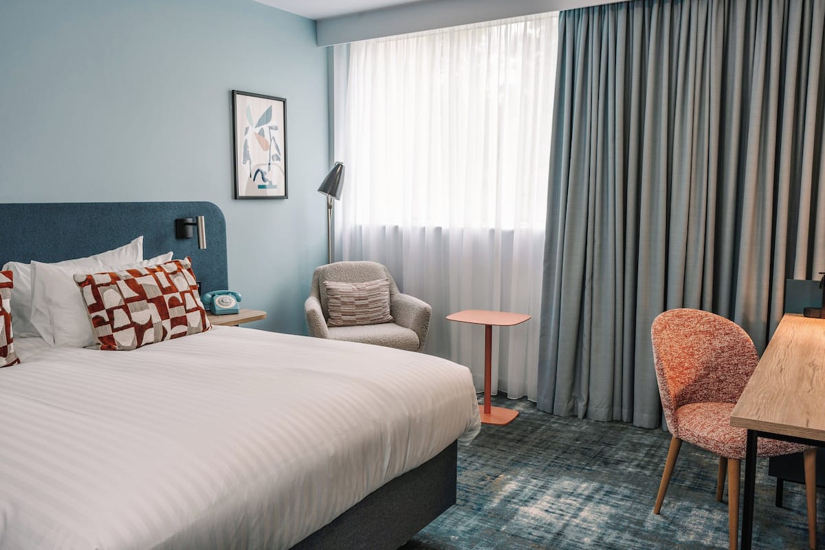 A hotel room with a large bed, a chair, a small table, and a window with curtains.