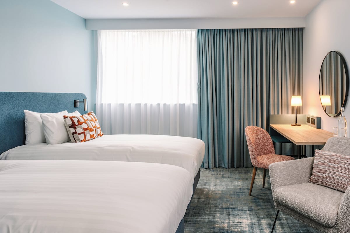 A hotel room with two beds, a desk, a chair, a mirror, and a view of the outdoors through a window with curtains.