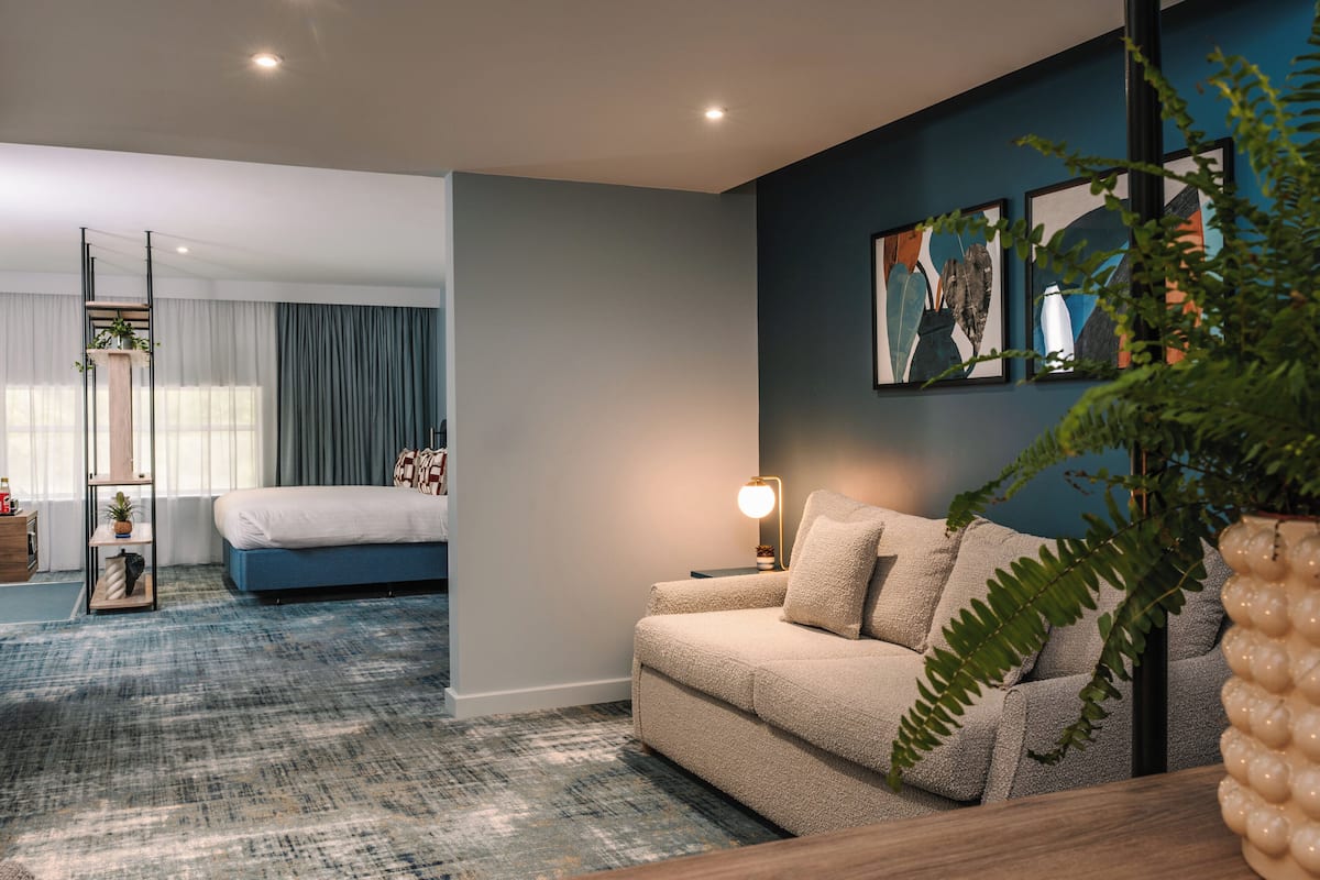 A modern hotel room with a large bed, a sofa, and a desk. There is a window with curtains and a plant on the desk.