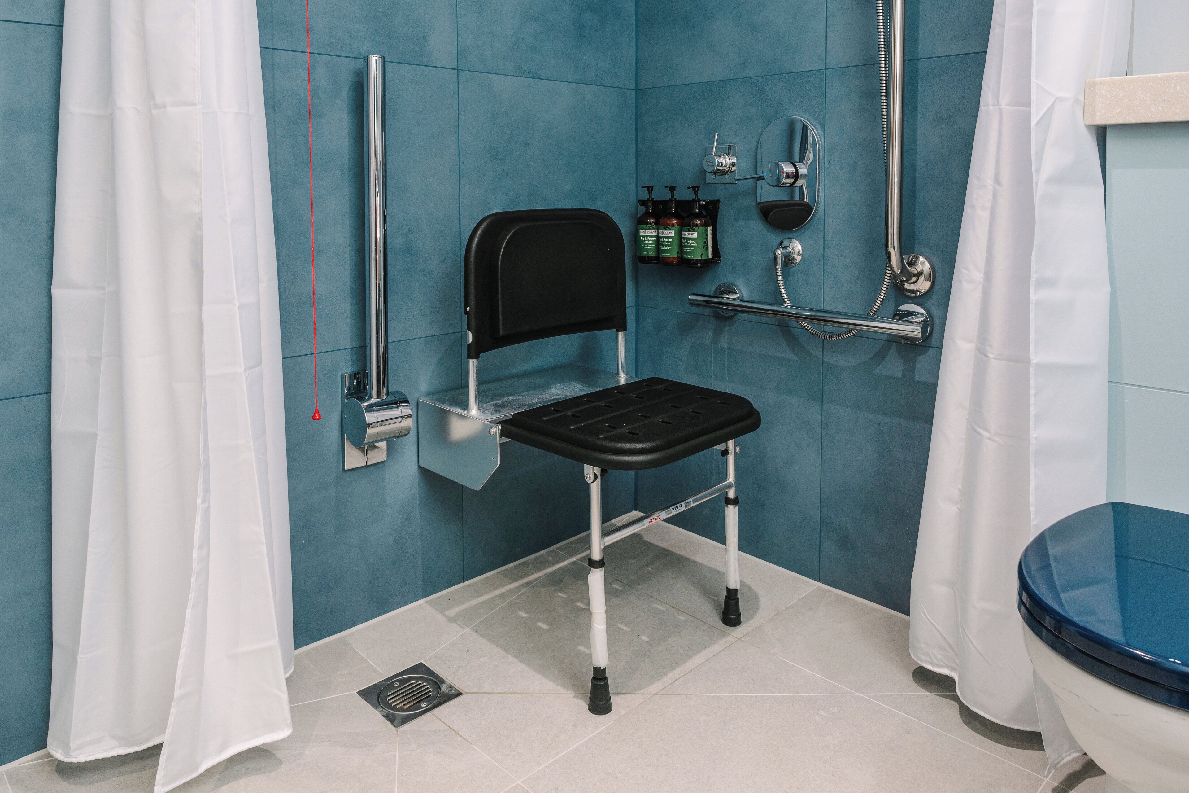 Un baño con ducha accesible para sillas de ruedas, barra de apoyo y retrete con asiento azul.