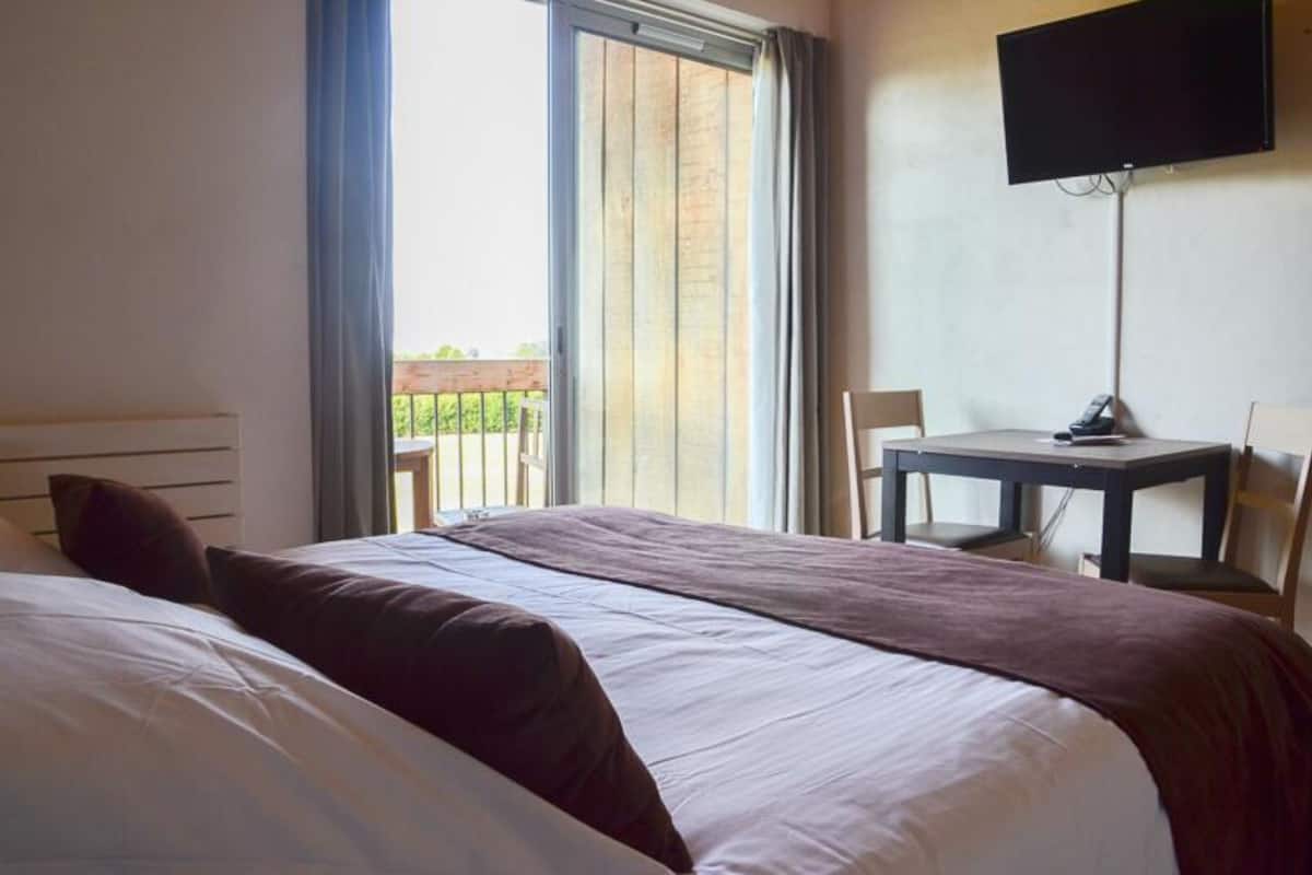 A hotel room with a large bed, a desk, a chair, a TV, and a balcony with a view.