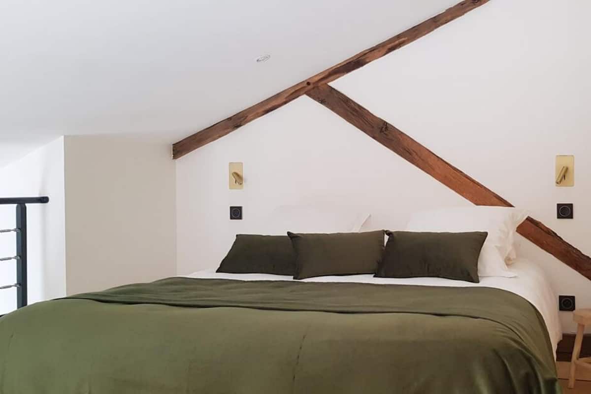 A bedroom with a bed, wooden beams, two wall-mounted light switches, and a small stool.