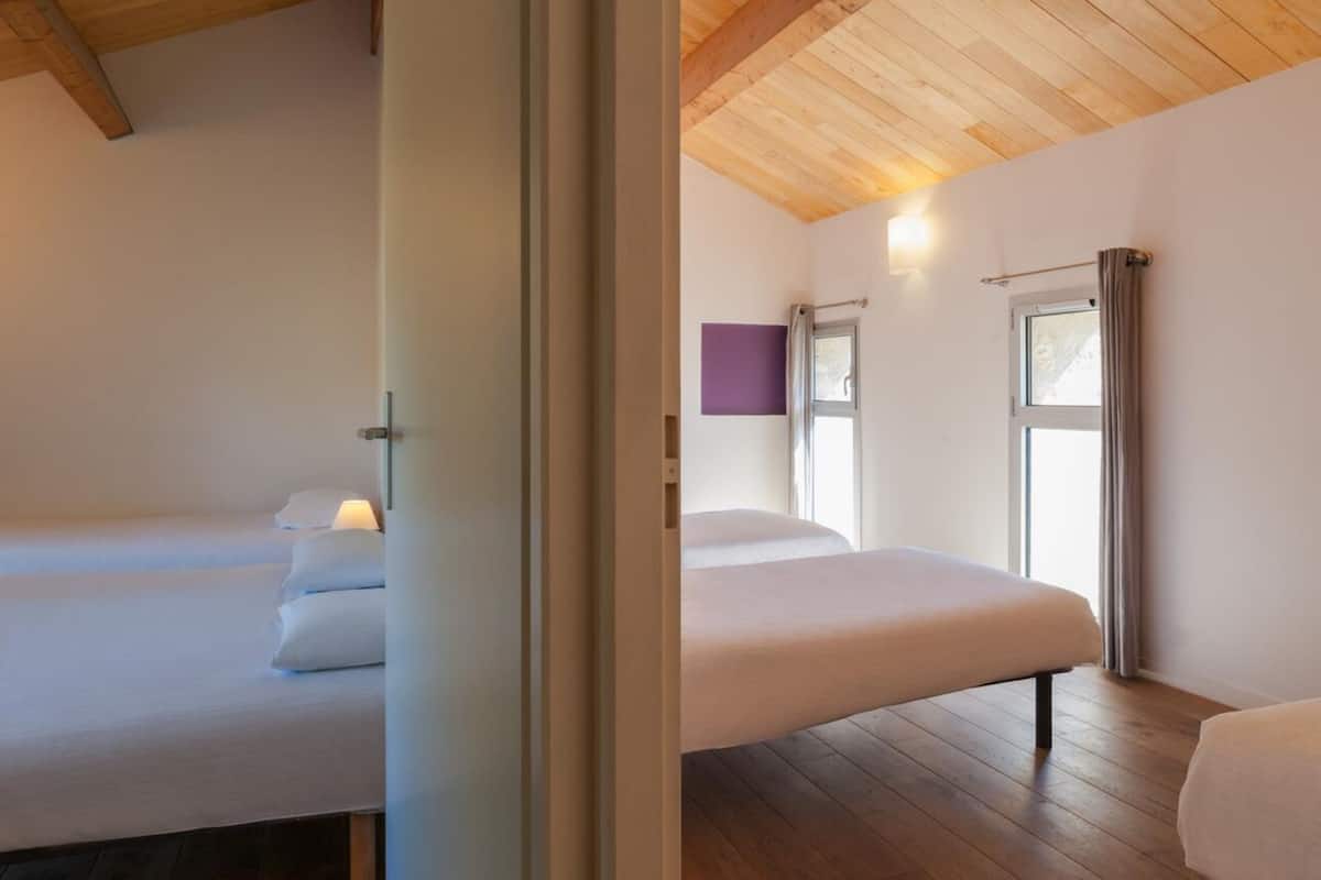 A hotel room with two beds, wooden flooring, a wooden ceiling, a window with curtains, and a wall-mounted light fixture.