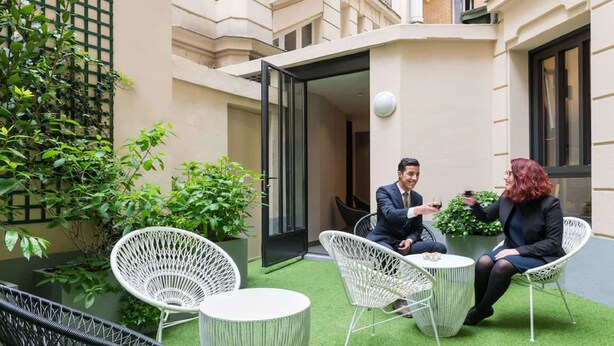 Un área de estar al aire libre con sillas de mimbre blanco y una pequeña mesa redonda, rodeada de plantas verdes y la entrada de un edificio.