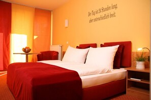Habitación de hotel con una cama grande, ropa de cama roja, una pequeña mesa con un cuenco de fruta y una pared con una cita en alemán.