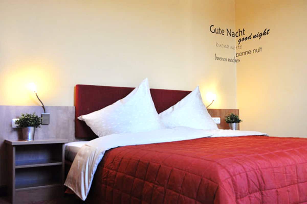 A neatly made bed with a red quilt, two bedside lamps, and a wall with a multilingual 'good night' message.