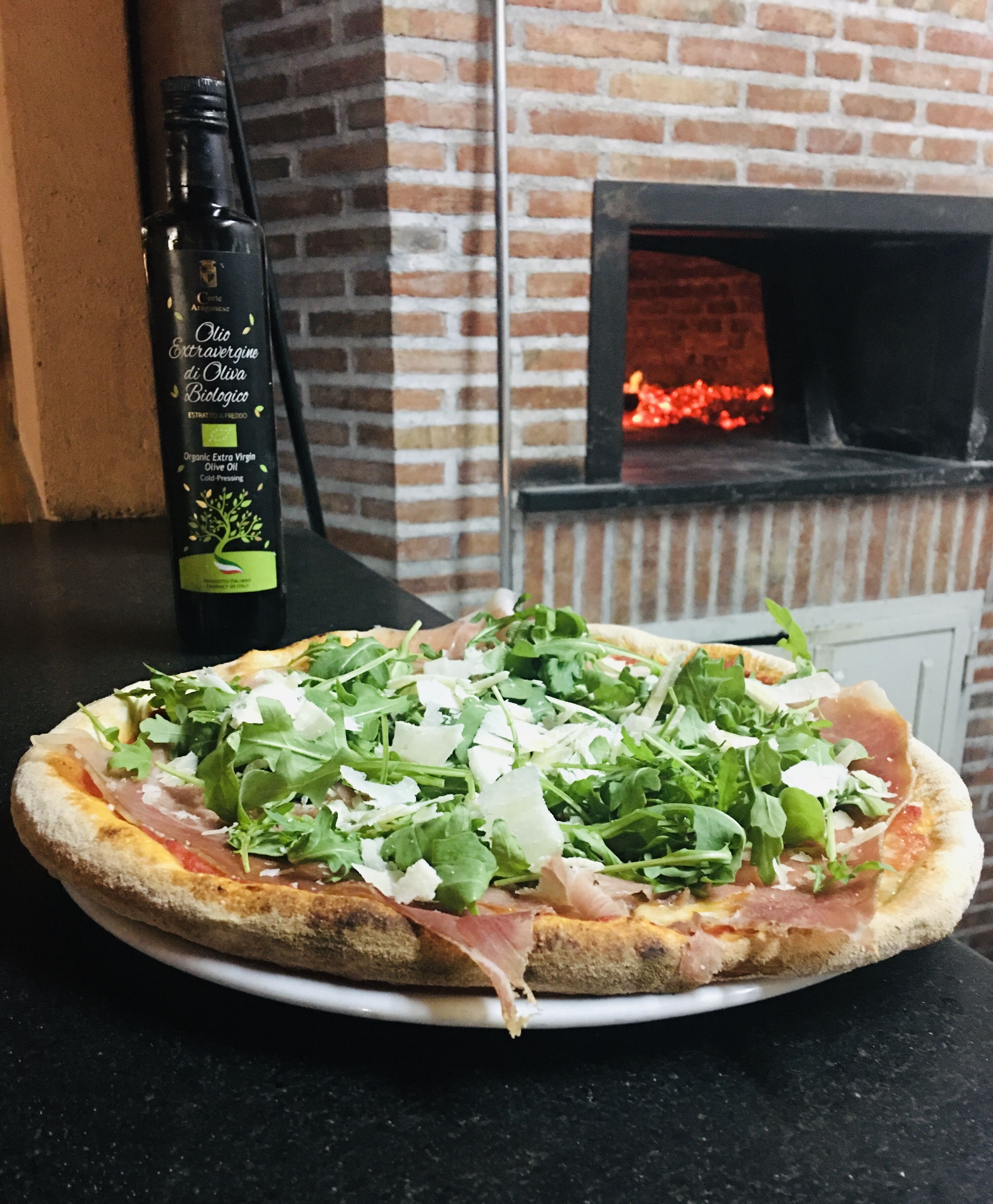 Pizza mit Gemüse und Käse auf einem Teller, eine Flasche Olivenöl und ein Steinofen im Hintergrund.