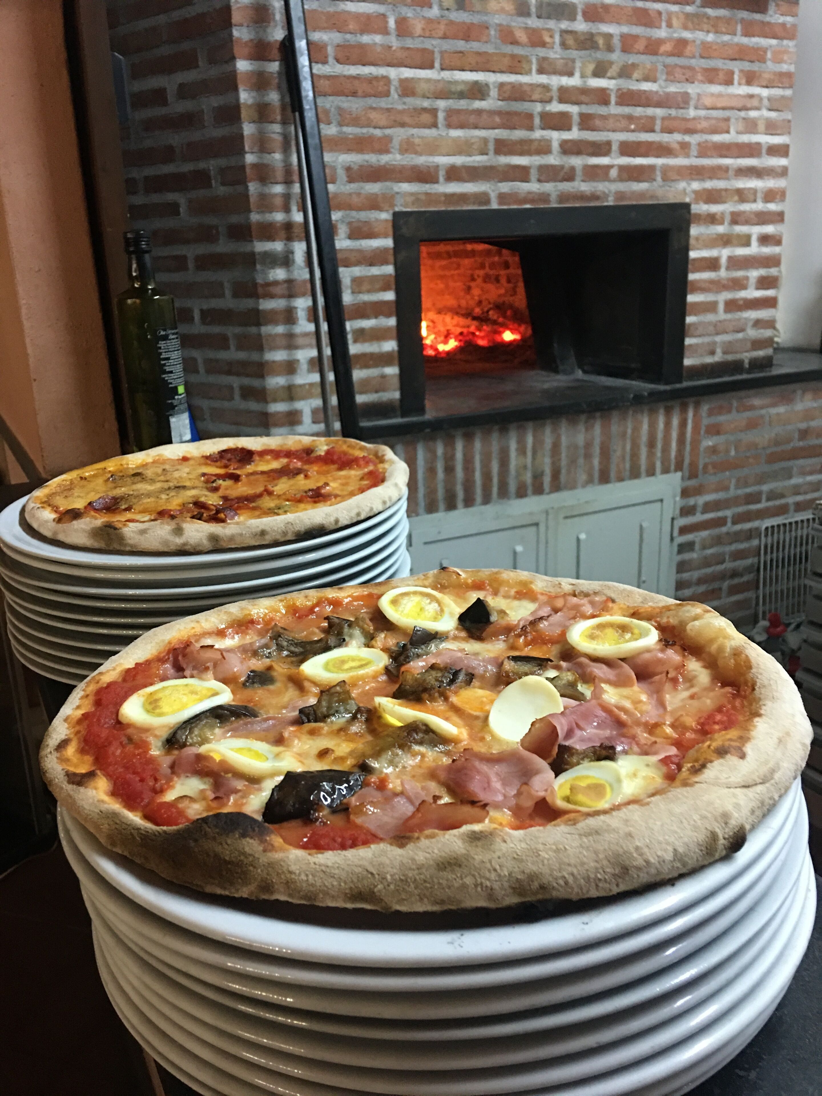 Dos pizzas sobre platos apilados frente a un horno de ladrillo.