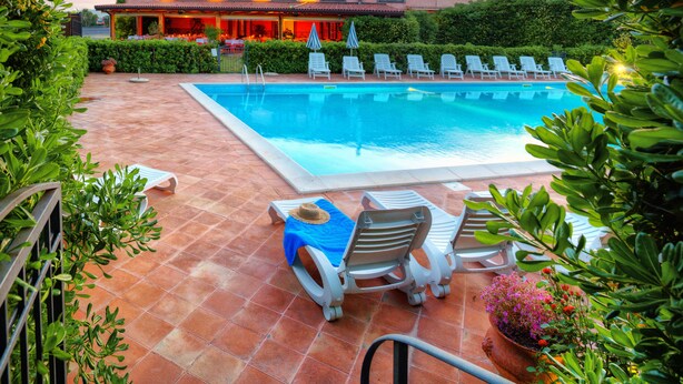 A swimming pool with lounge chairs, a tiled patio, and a building with a red roof in the background. - Corte Aragonese (Santa Maria di Licodia)