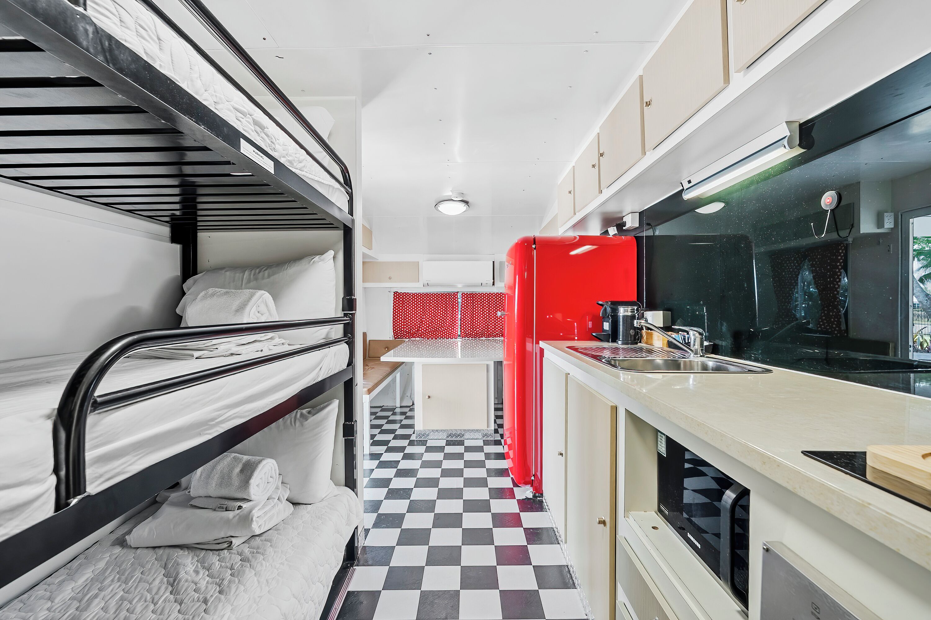 Une pièce compacte avec des lits superposés, un coin cuisine, un sol à carreaux et un mur d’accent rouge.