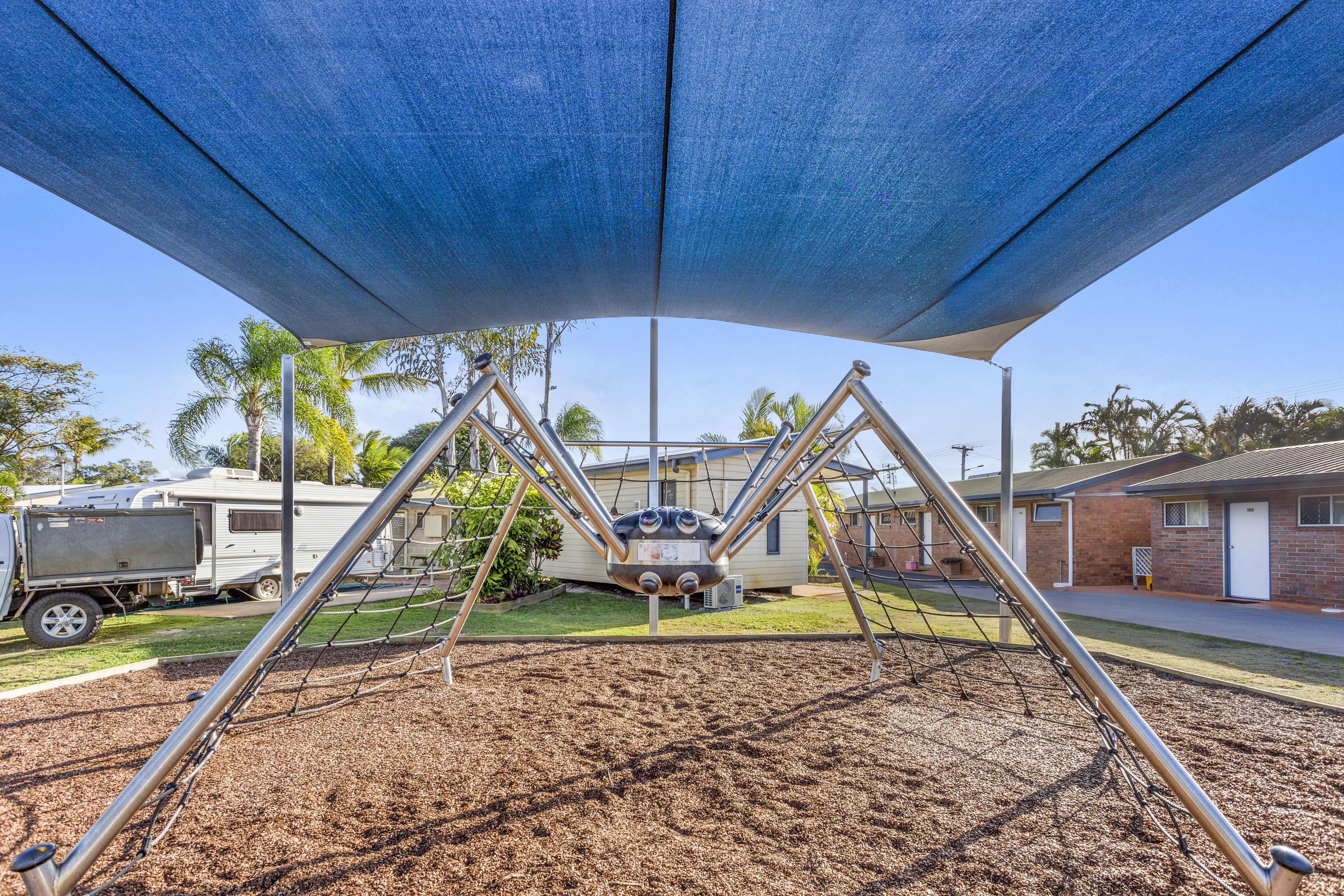 Un parque infantil con columpios bajo un toldo azul, rodeado de una zona residencial con casas y una caravana.