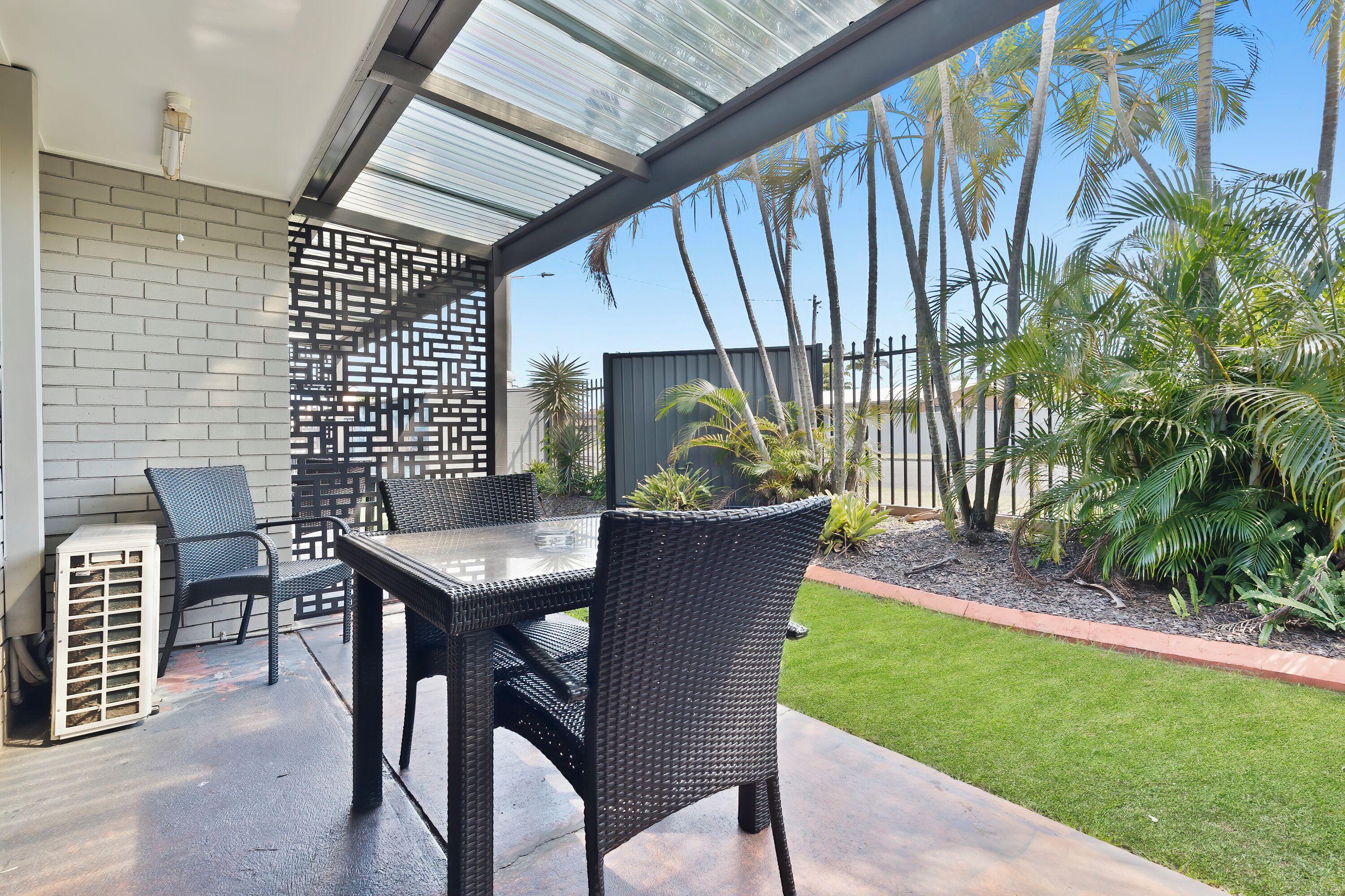 Une terrasse extérieure avec une table et des chaises, une véranda fermée par des vitres, et une vue sur des palmiers et une clôture.