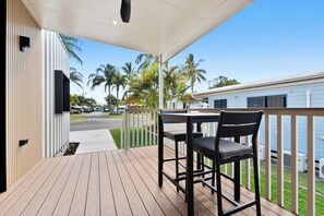 A wooden deck with a table and chairs, overlooking a street with palm trees and a blue building. - Ingenia Holidays Hervey Bay (Torquay)