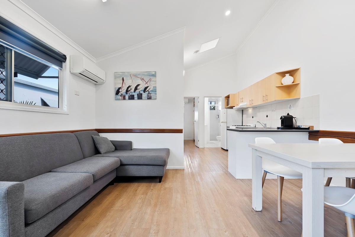 Ein modernes Wohnzimmer mit einer grauen Eckcouch, einem Esstisch mit Stühlen, einer Küchenzeile mit Holzschränken und einem großen Fenster mit Aussicht.