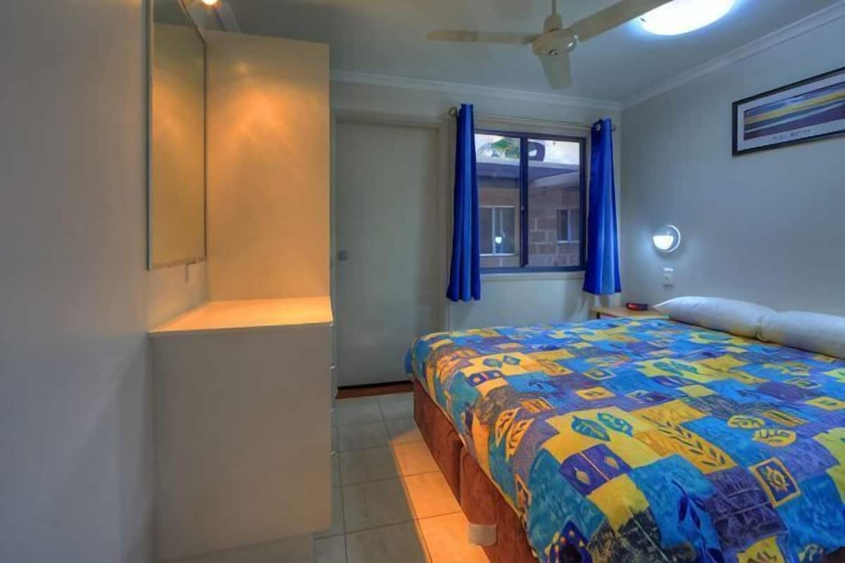 Ein Schlafzimmer mit einem Bett, einem Deckenventilator, einem Fenster mit Vorhängen und einer Wandleuchte.