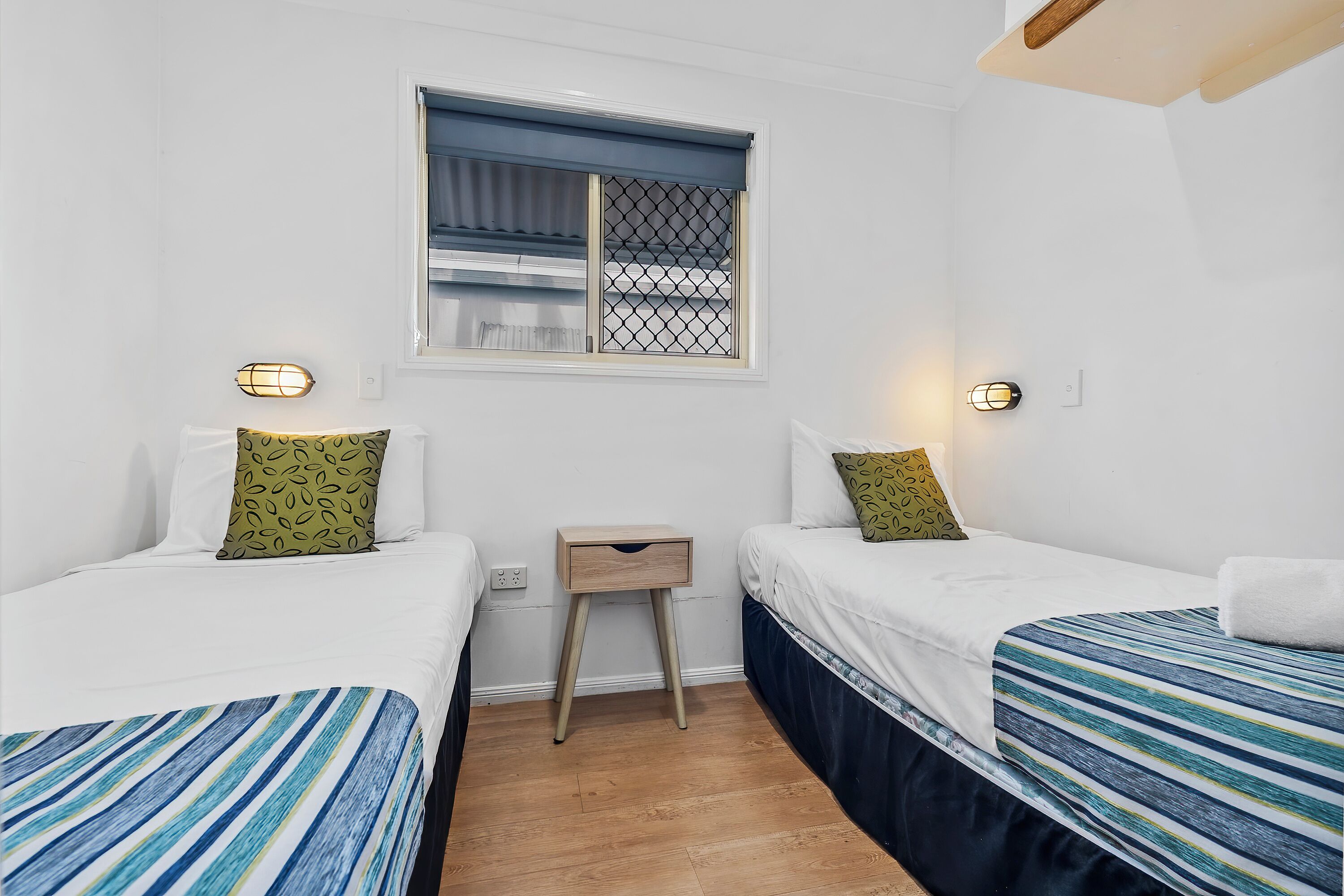 Ein Zimmer mit zwei Betten, einem kleinen Tisch, einem Fenster mit Jalousie und Wandleuchten.