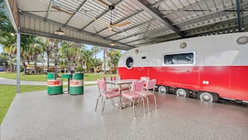 A covered outdoor area with a table and chairs, a red and white train, and green trash bins.