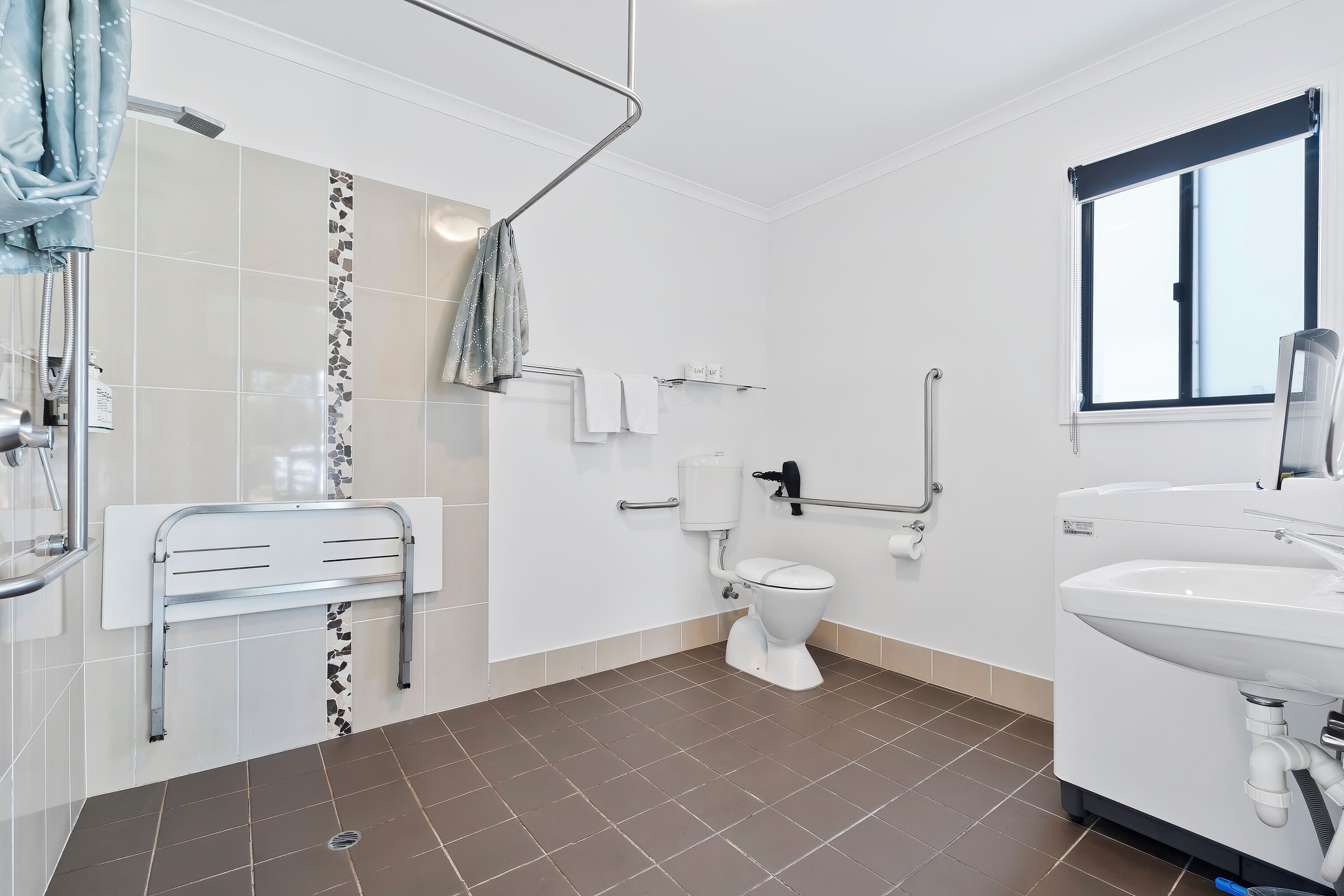 Ein Badezimmer mit Dusche, Toilette und Waschbecken, ausgestattet mit Haltegriffen und einem Fenster.