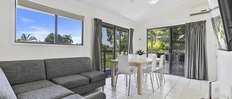 A modern living room with a grey sofa, a dining area with a table and chairs, and large windows with curtains.