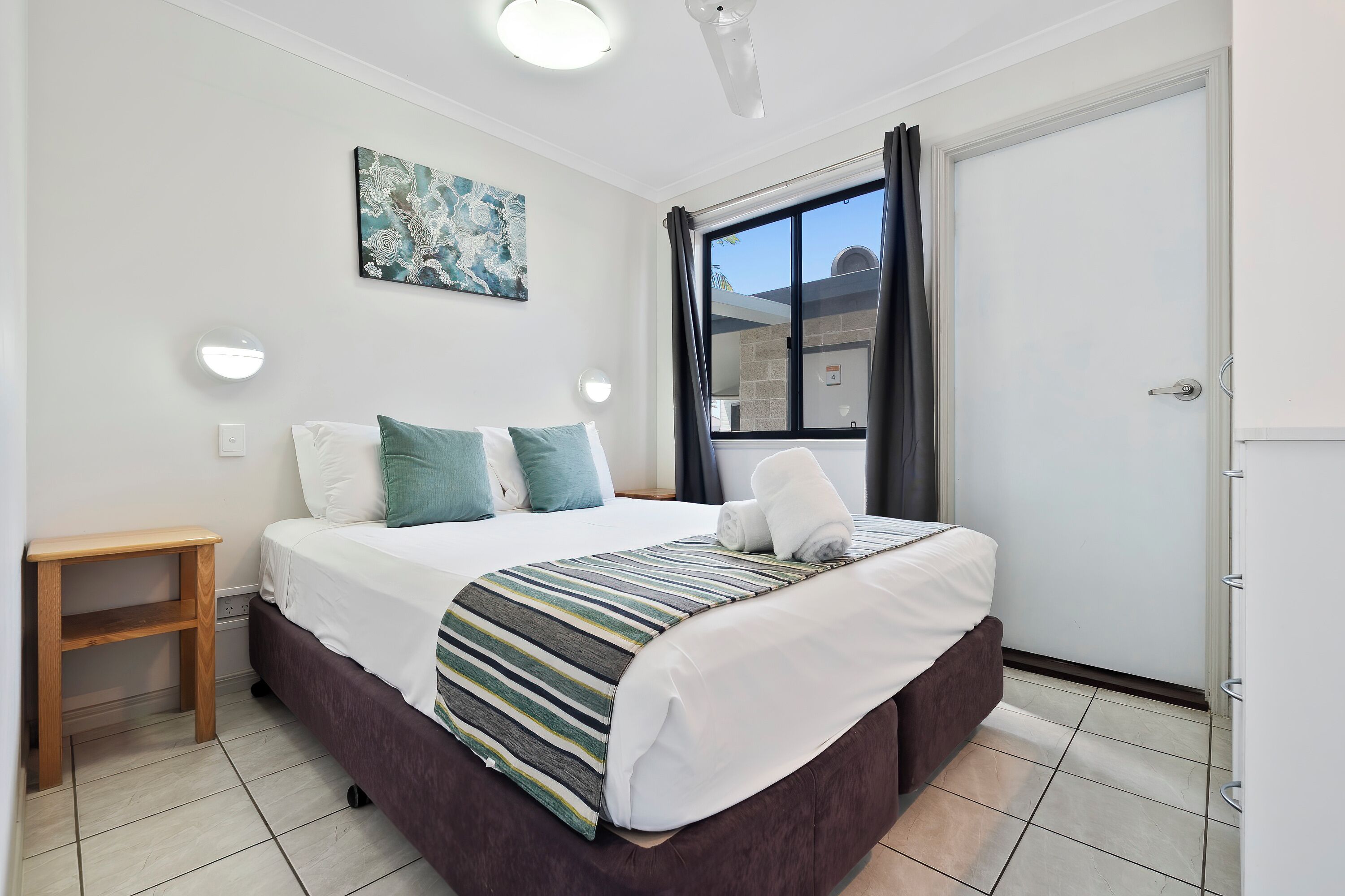 A neatly made bed with a striped blanket, a wooden nightstand, a wall-mounted light, and a window with curtains.