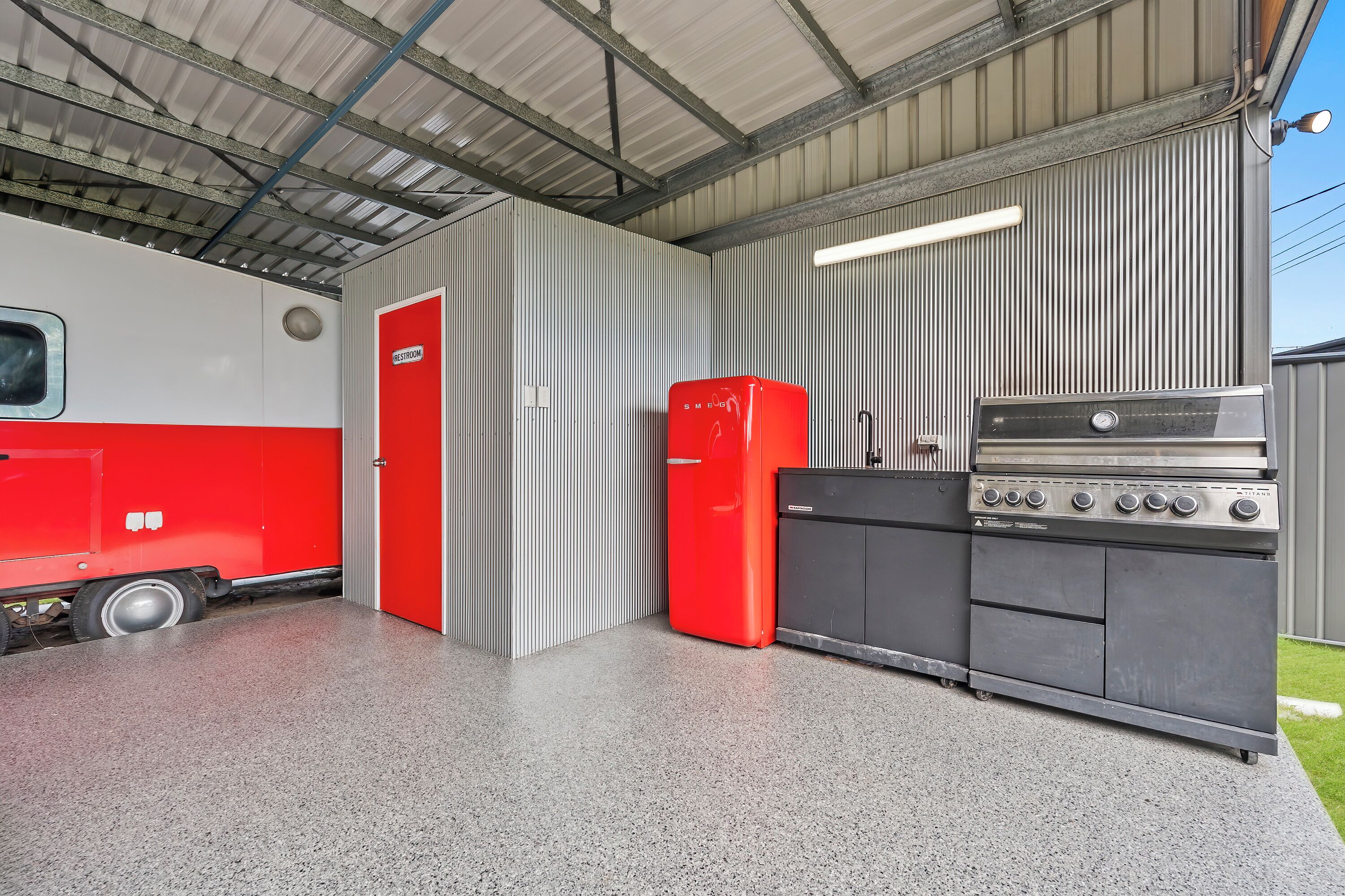 Zona de cuina amb un frigorífic vermell, un forn d'acer inoxidable i una porta amb la inscripció 'RESTROOM'.