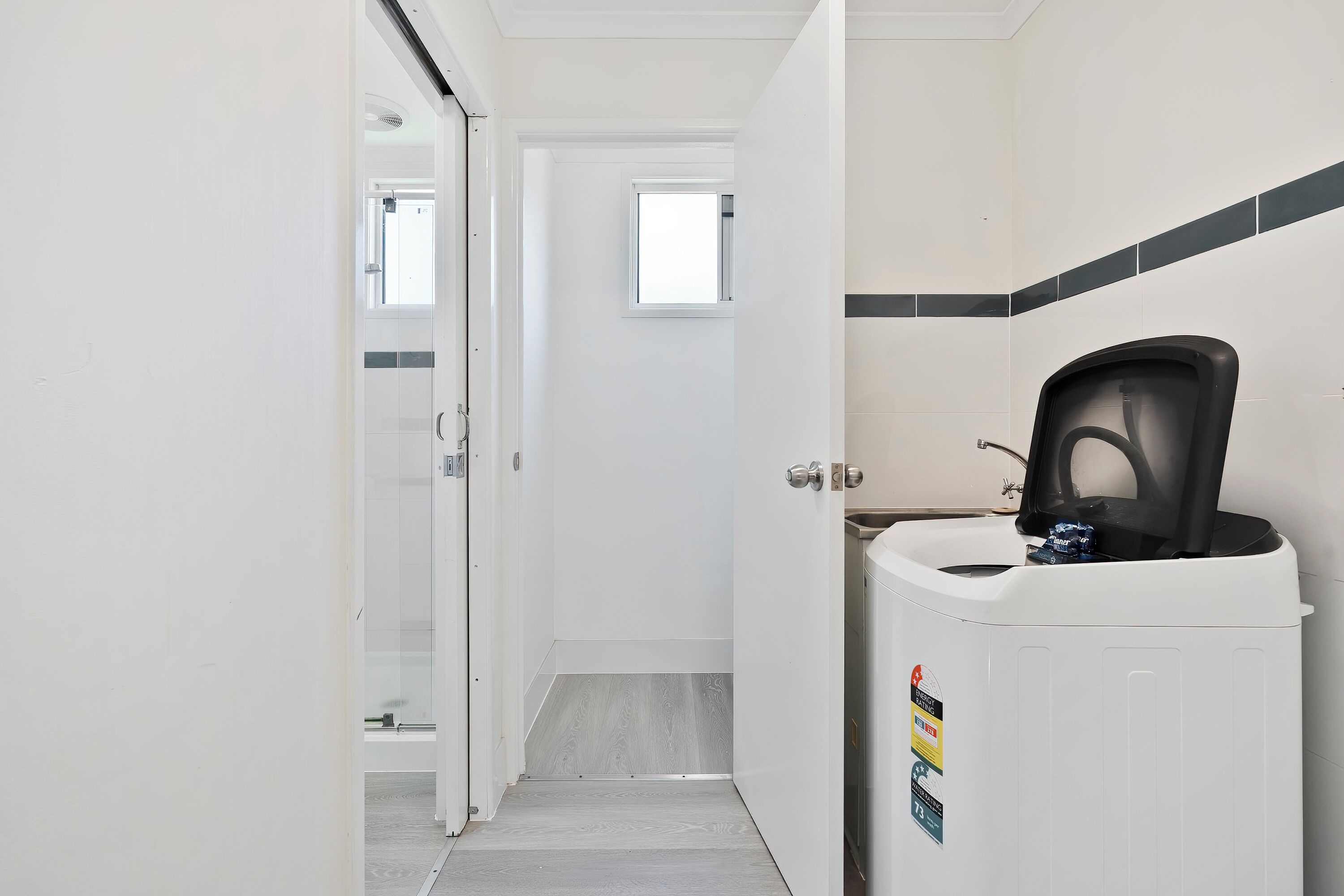 A narrow hallway with a white door at the end, a small window, and a coffee machine on the right.