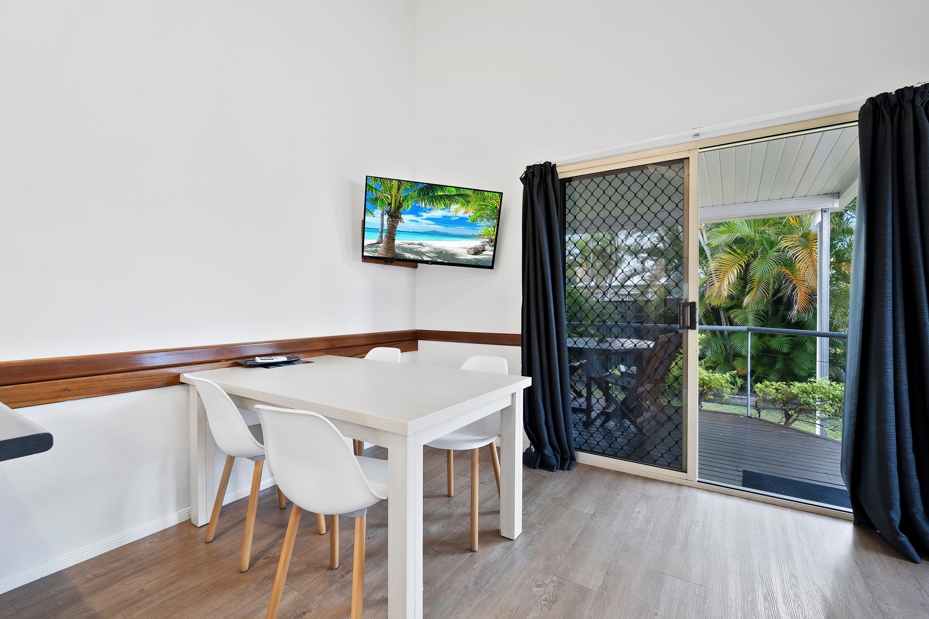 Una habitación con una mesa y sillas blancas, un televisor en la pared y un ventanal corredero a un balcón con vistas a zonas verdes.