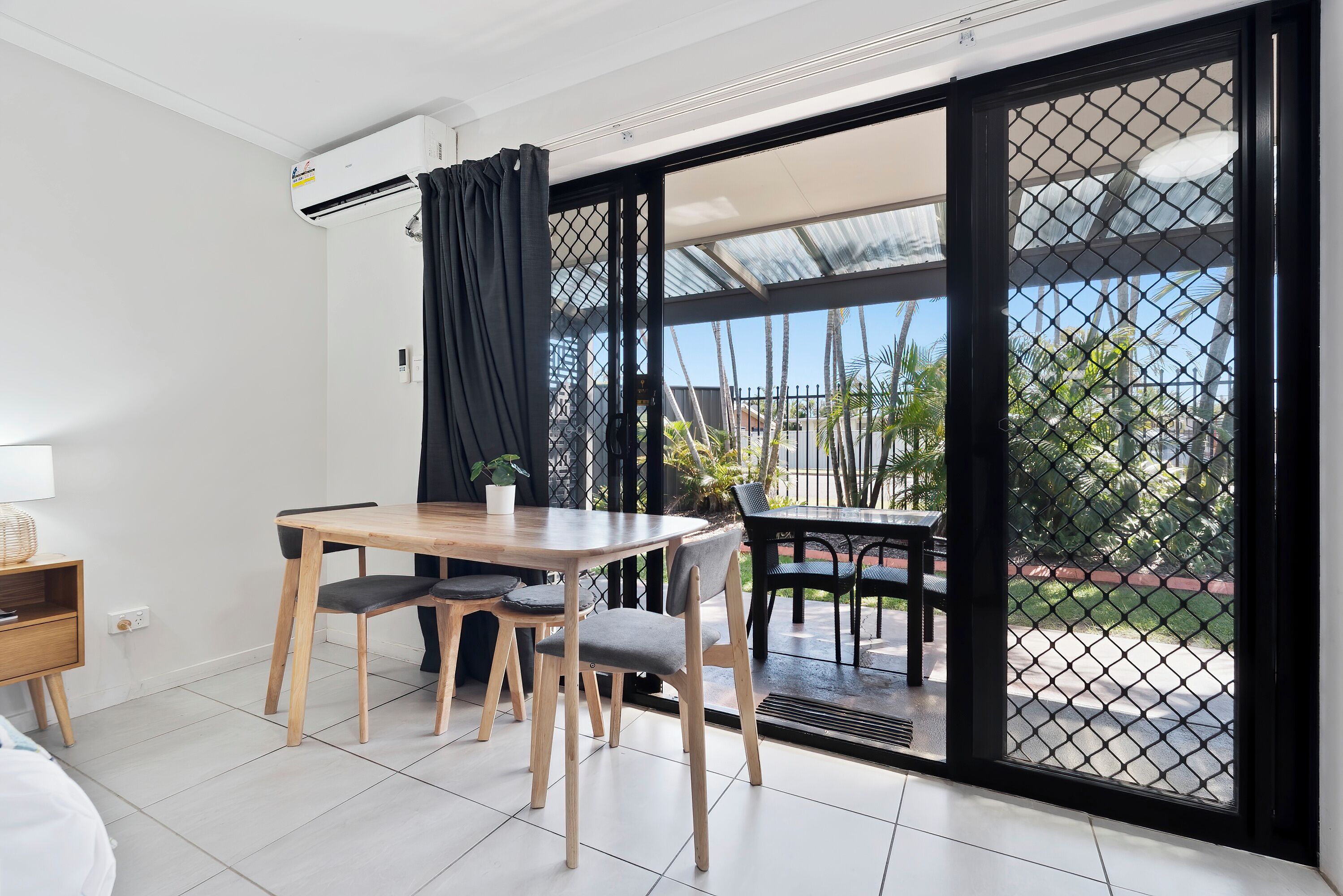 A room with a dining table, chairs, a small plant, and a sliding glass door leading to an outdoor area with a fence and greenery.