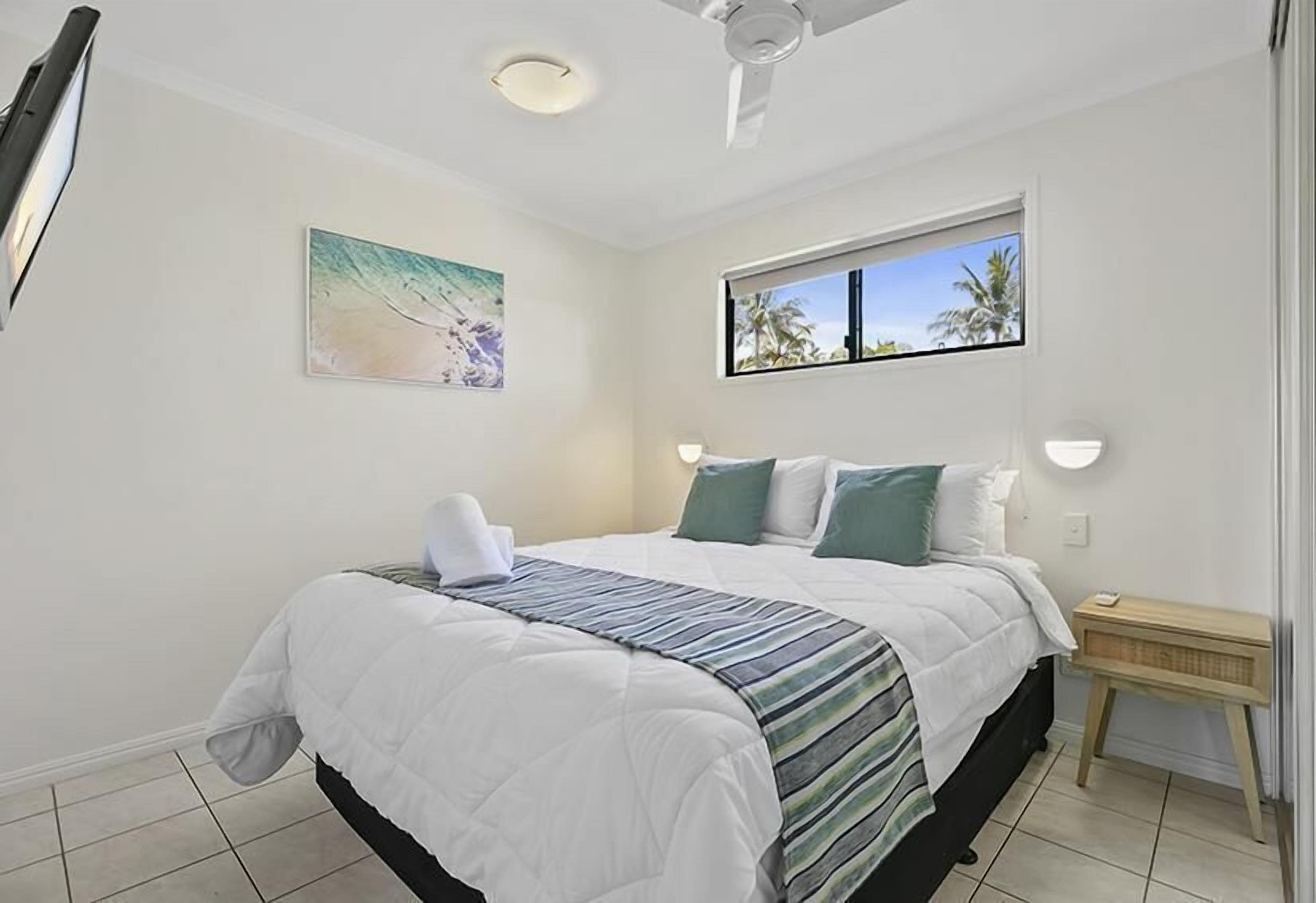 Un dormitorio con cama, ventilador de techo, ventana con vistas a palmeras y un cuadro en la pared.