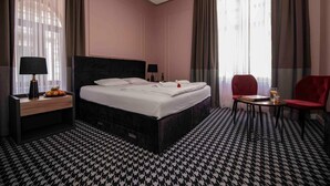 Ein Hotelzimmer mit einem großen Bett, zwei Nachttischen, einem kleinen Tisch mit einem Stuhl und einem Fenster mit Vorhängen.