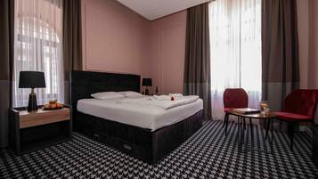 Ein Hotelzimmer mit einem großen Bett, zwei Nachttischen inklusive Lampen, einem kleinen Tisch mit einem Stuhl und Blick ins Freie durch das Fenster.