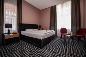 Ein Hotelzimmer mit einem großen Bett, zwei Nachttischen, einem kleinen Tisch mit einem Stuhl und einem Fenster mit Vorhängen.