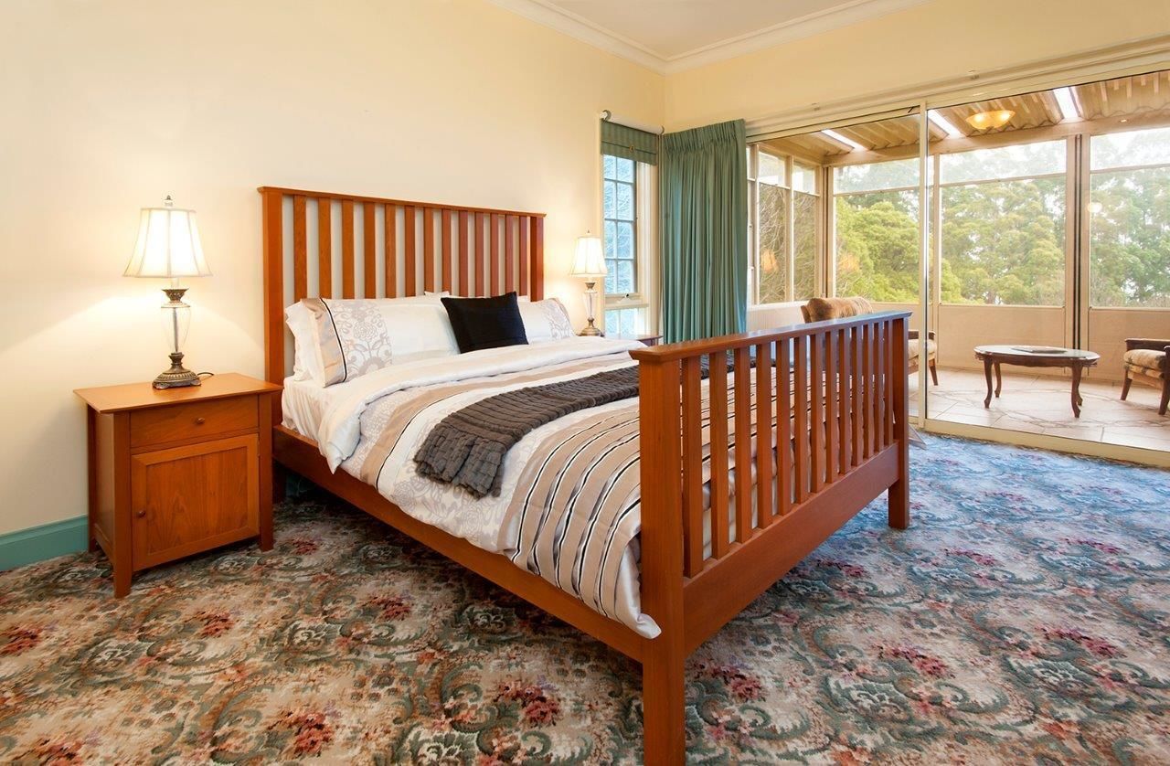 Quarto com cama de madeira, mesa de cabeceira, janela com cortinas e varanda com vista para árvores.