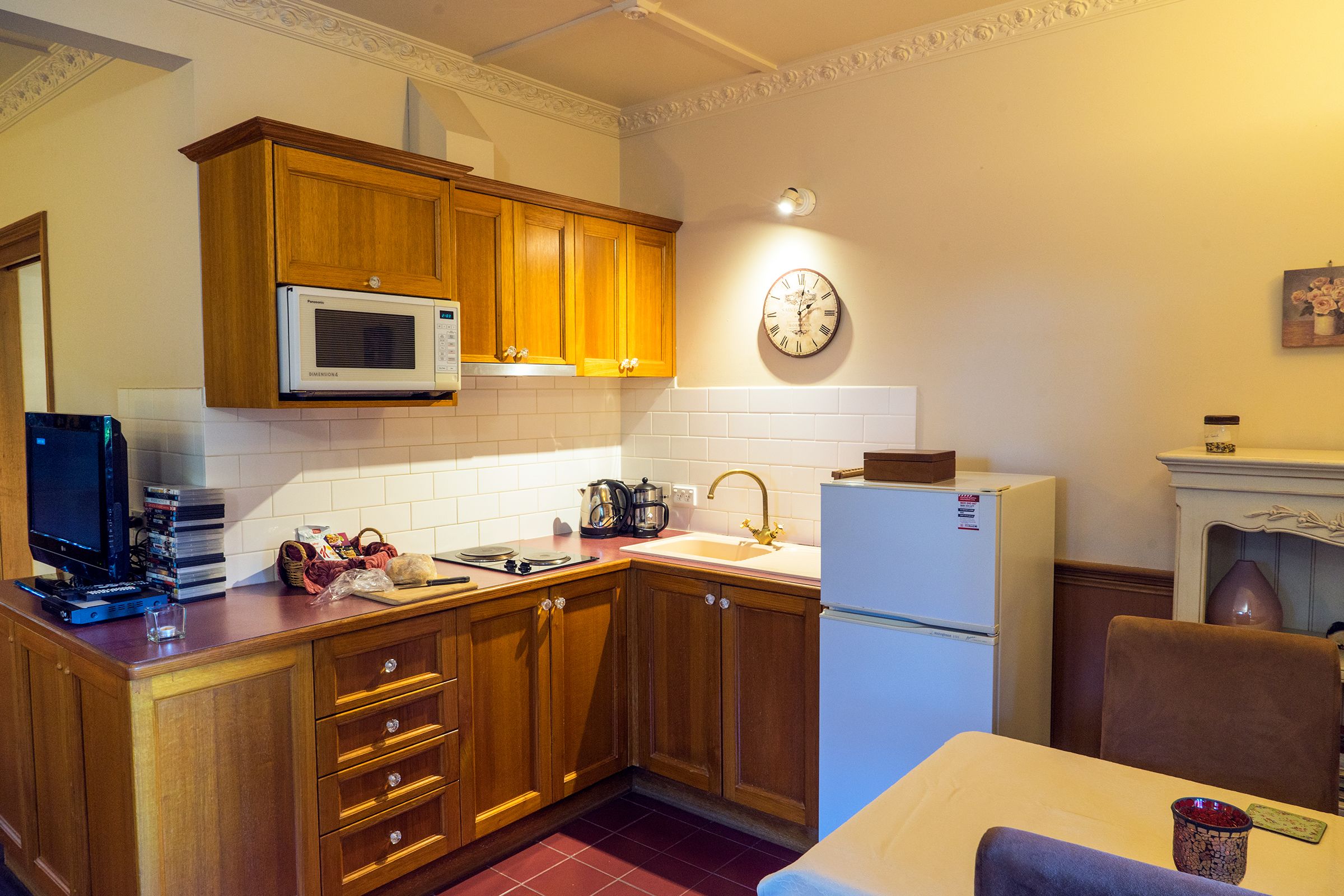 A compact kitchen with wooden cabinets, a microwave, a sink, and a clock on the wall.