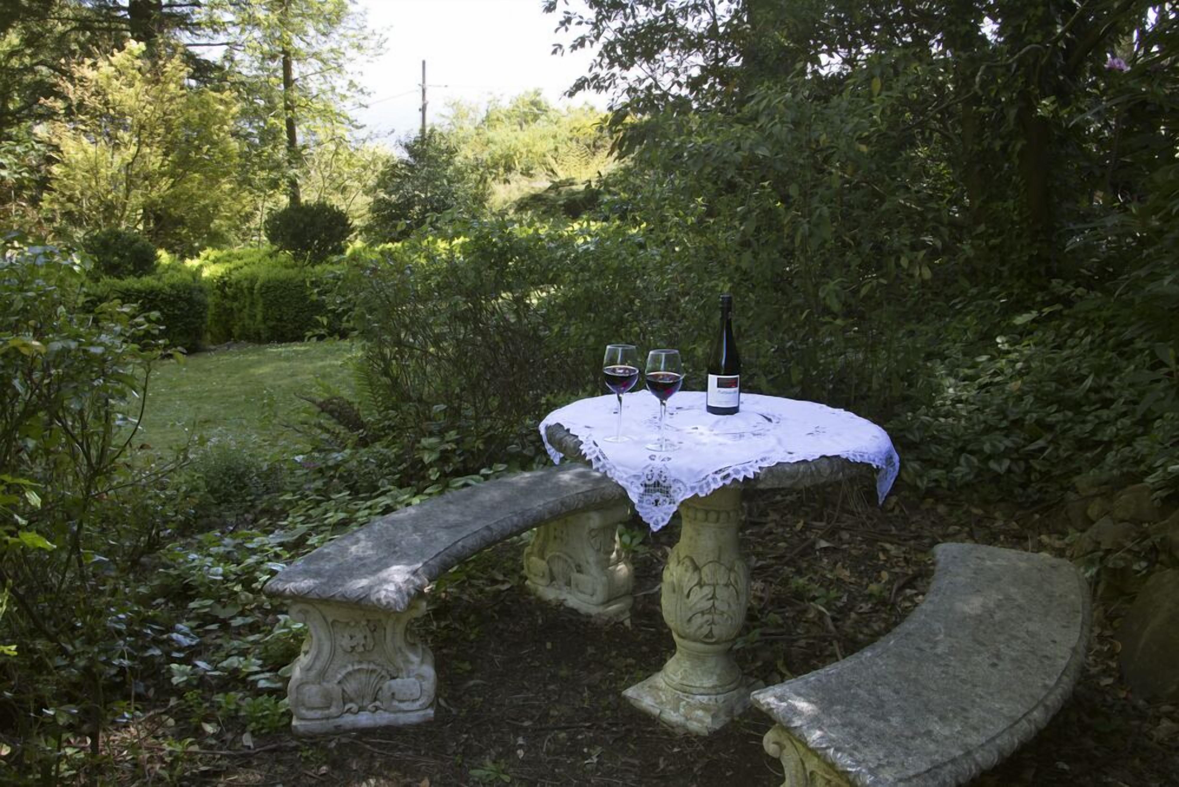 A stone bench and table set in a lush garden with a white lace tablecloth, two wine glasses, and a bottle.