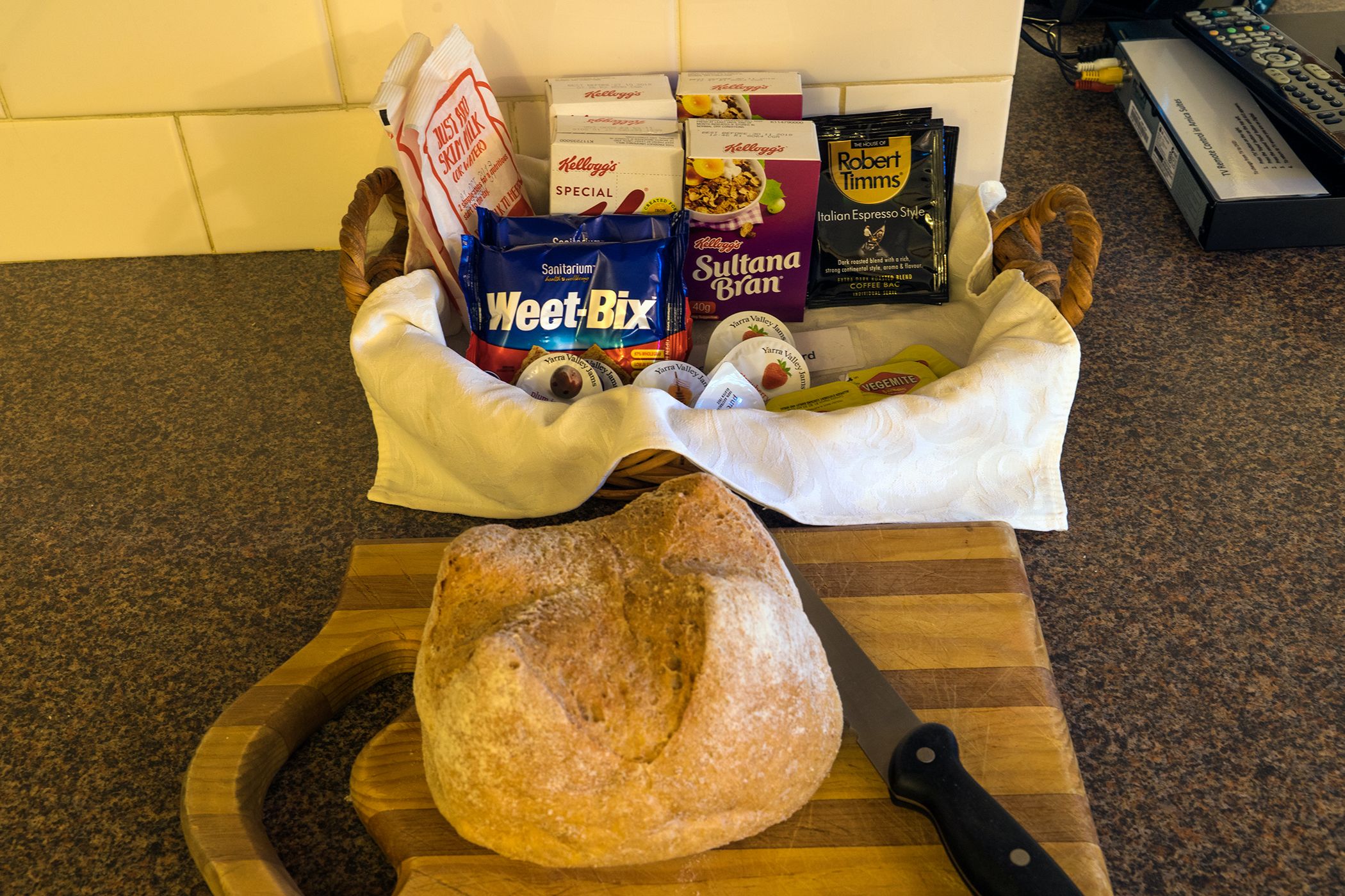 A loaf of bread on a wooden cutting board, a knife, and various packaged food items including Weet-Bix, Sultana Bran, and Italian Espresso Style coffee.