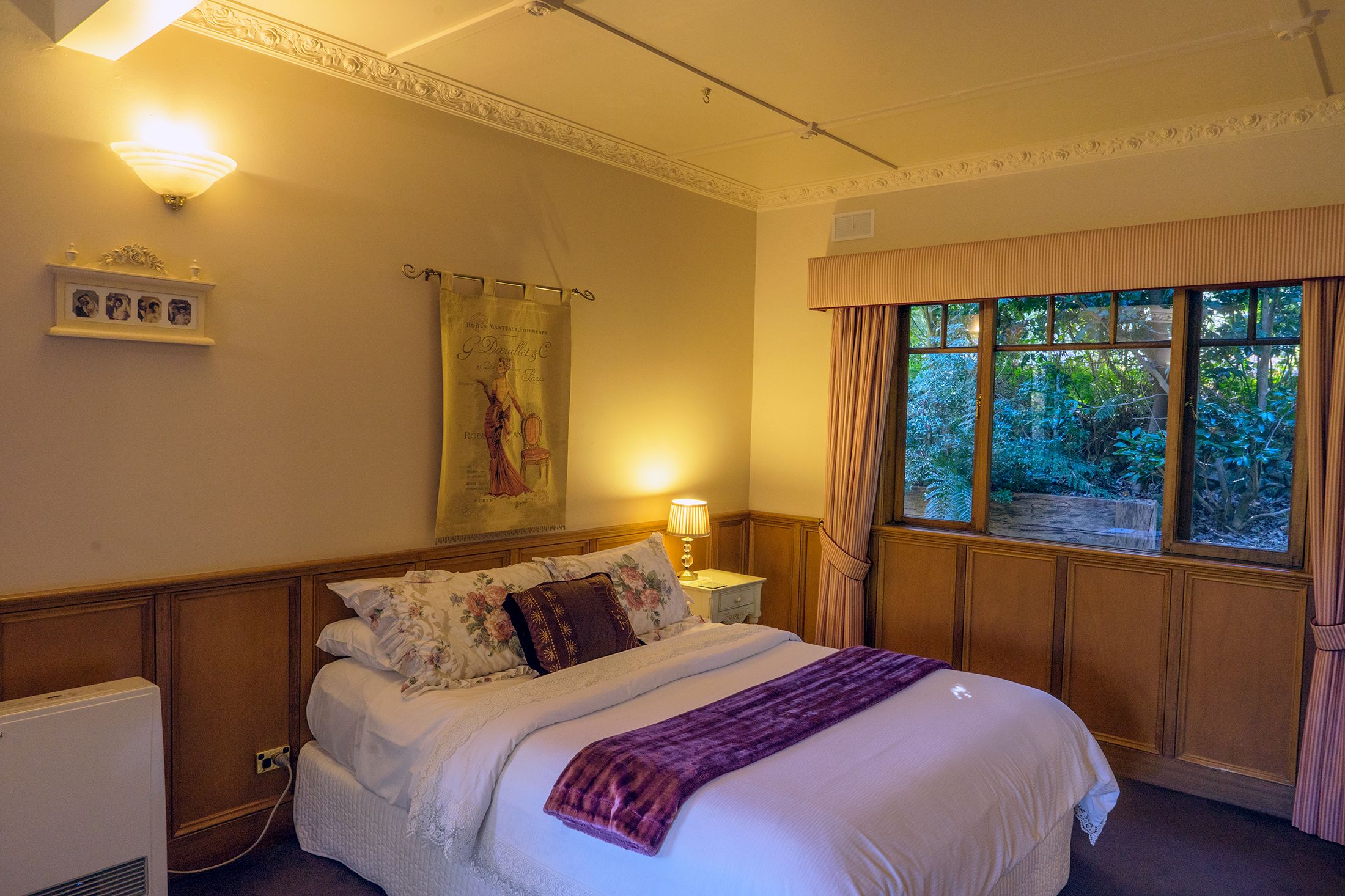 Una cama bien hecha con una manta morada, una mesita de noche con lámpara, un cuadro colgado en la pared y una ventana con cortinas.