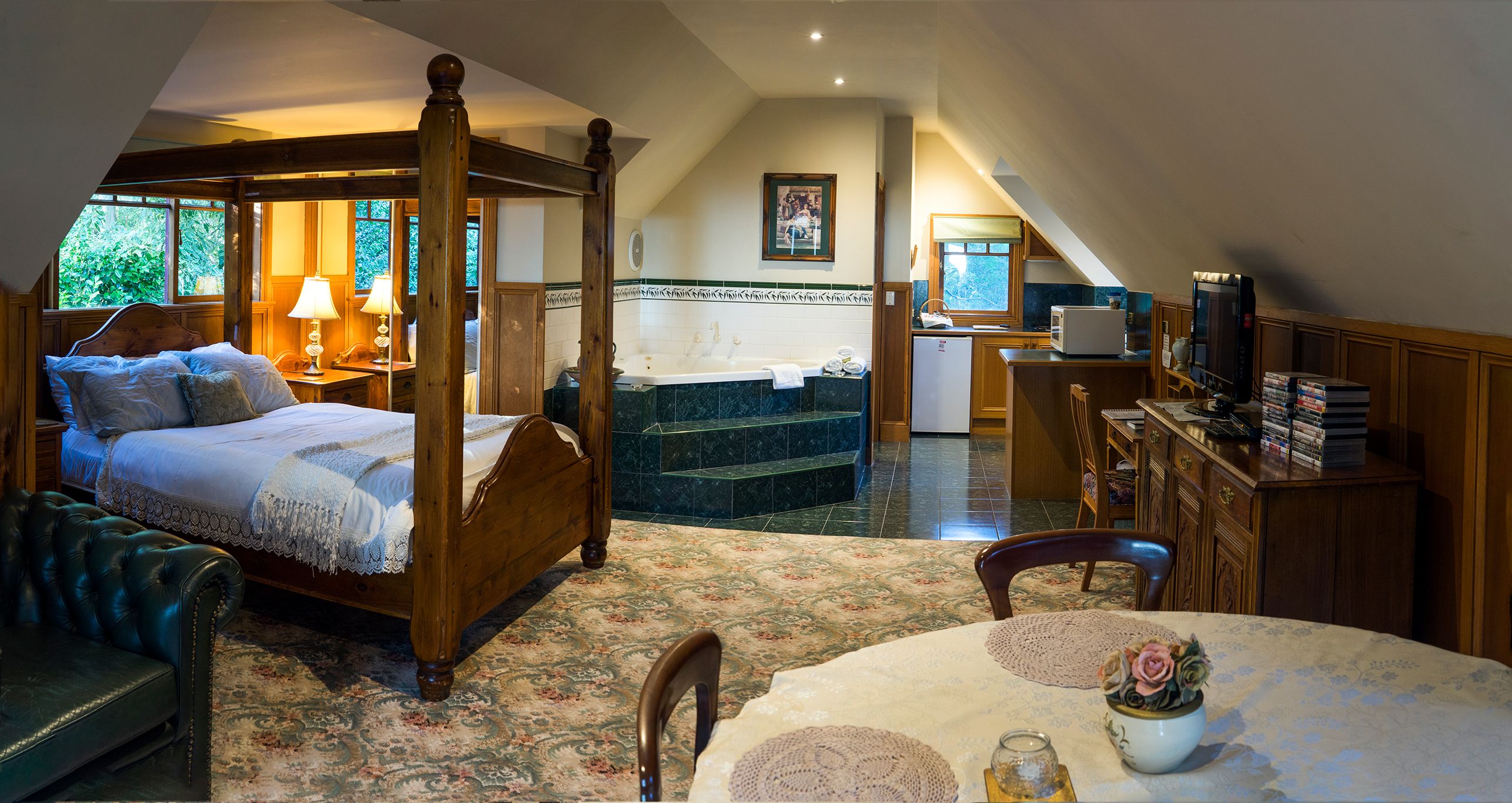 A bedroom with a four-poster bed, a sitting area with a sofa, a dining table with a vase of flowers, and a kitchen area with a refrigerator and books.