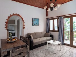 Una sala de estar acogedora con una mesa de madera, un sofá y una pequeña mesa blanca. Se ve una cocina al fondo, y hay una ventana con cortinas.