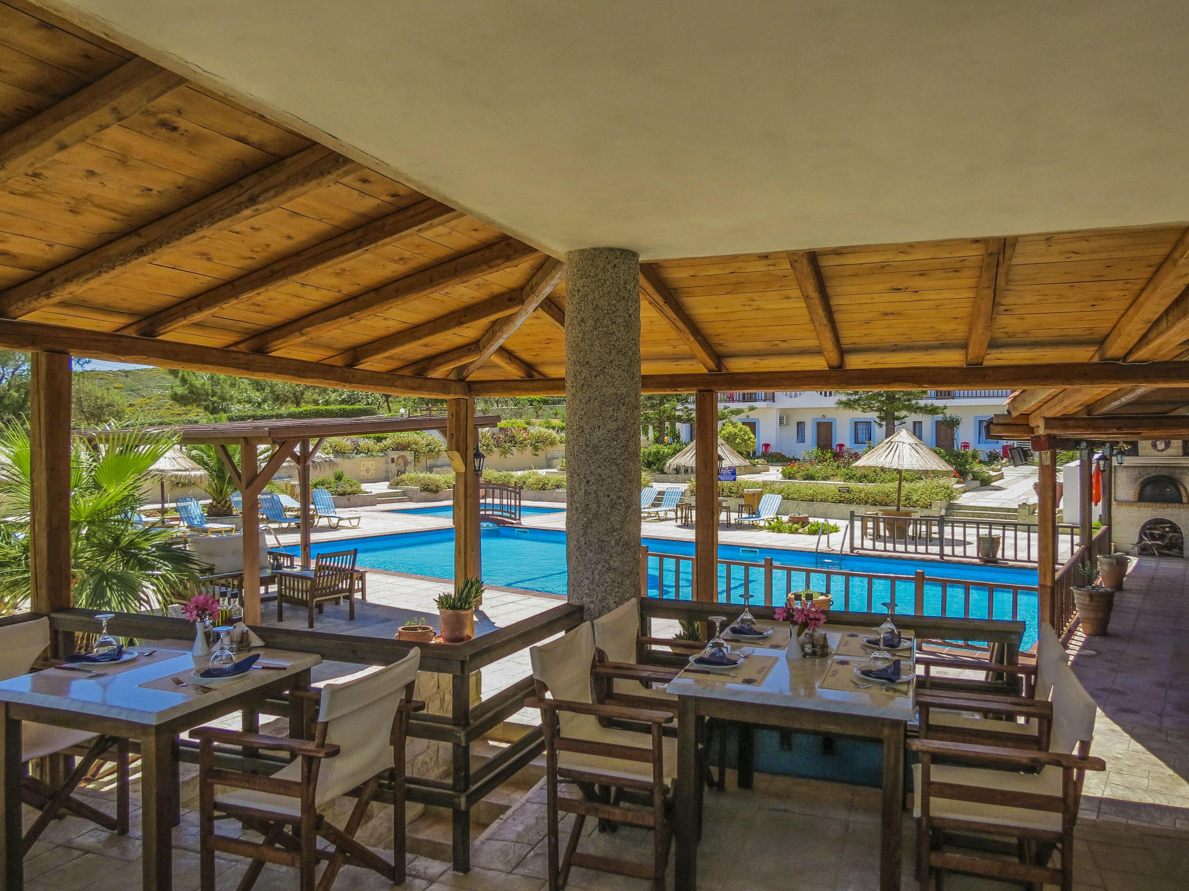 Un área de comedor al aire libre con techo, mesas y sillas, con vistas a una piscina y edificios al fondo.