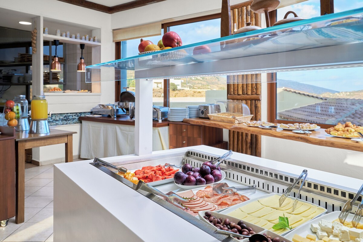 Un bufé con diversas opciones de comida, entre ellas frutas, quesos y carnes, en una sala bien iluminada con vistas al exterior.