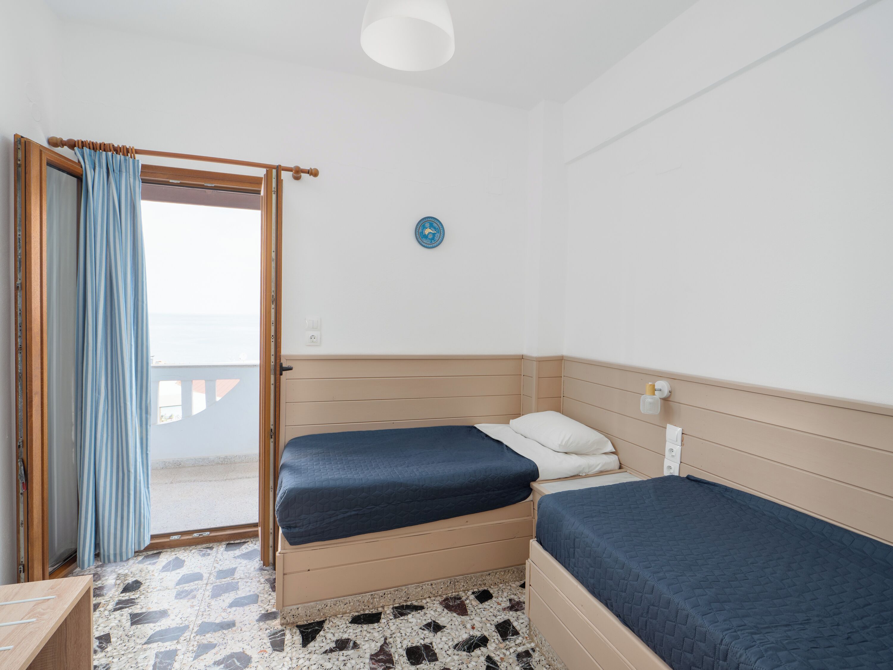 Una habitación con dos camas, cabecera de madera, pared blanca, un reloj redondo pequeño y una ventana con cortina.