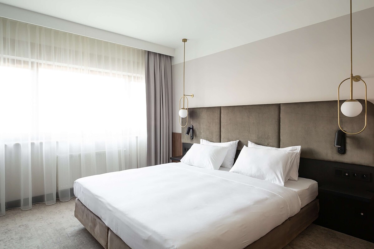 Une chambre d’hôtel avec un grand lit, une tête de lit, une table de chevet et une fenêtre avec des rideaux.