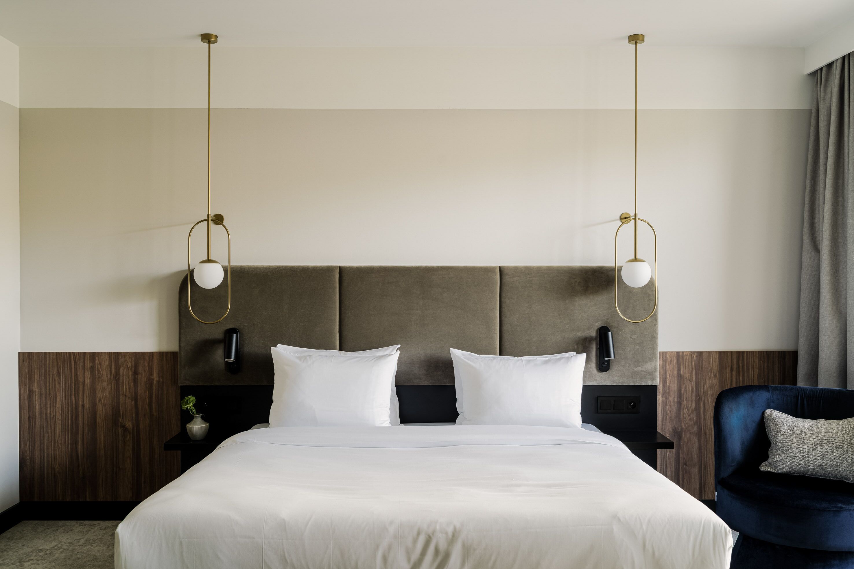 Quarto de hotel com uma cama grande, duas mesas de cabeceira, uma poltrona azul escura e uma cabeceira moderna.