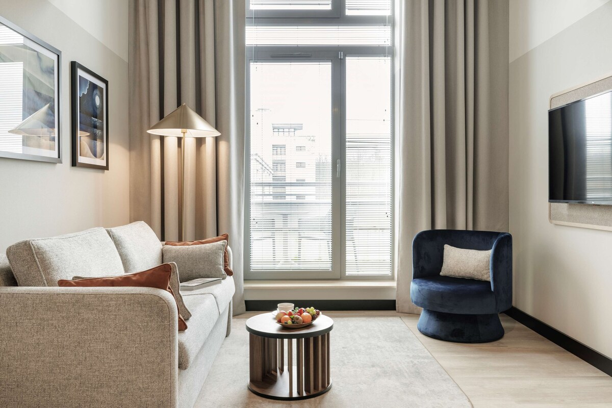 Un salon moderne avec un canapé, une table basse ronde sur laquelle sont disposés des fruits, un fauteuil bleu et une grande fenêtre avec des stores.